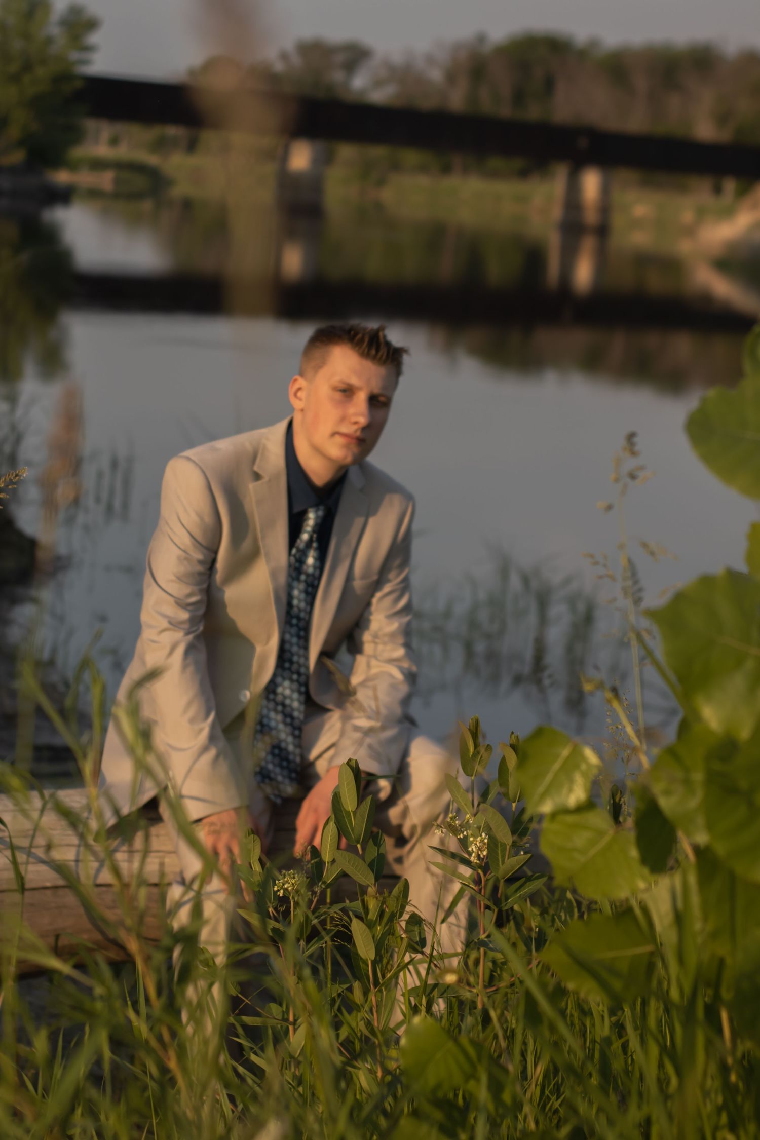 Person in formal attire seated among tall grass near a tranquil pond at sunset.