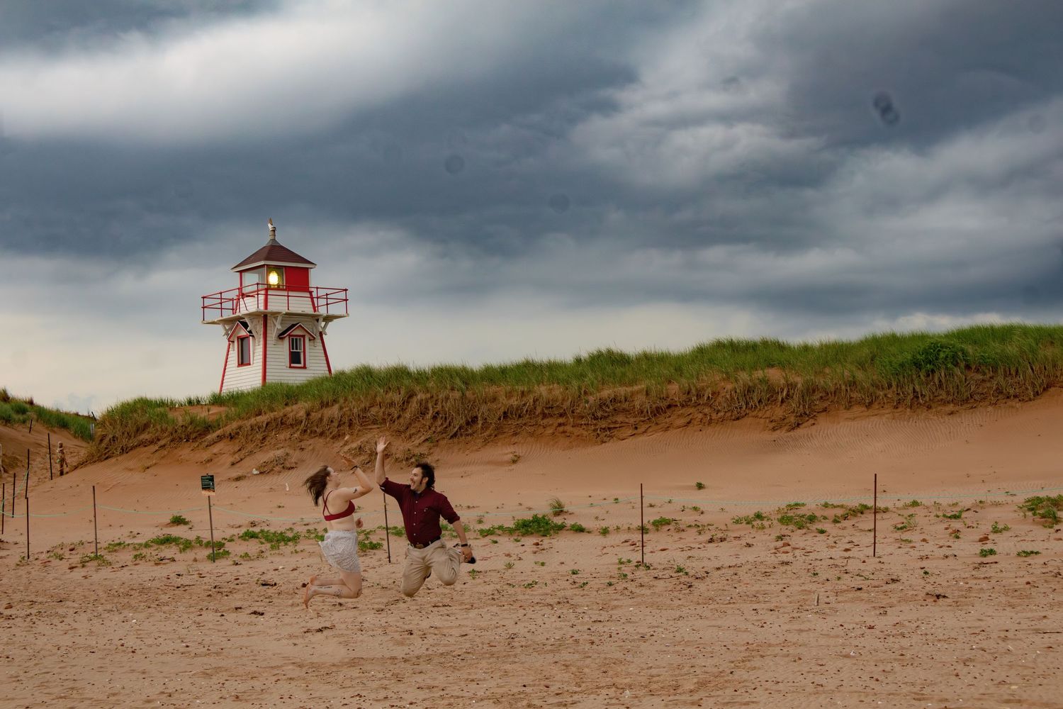 A red and white lighthouse perches atop sandy dunes against moody sky.