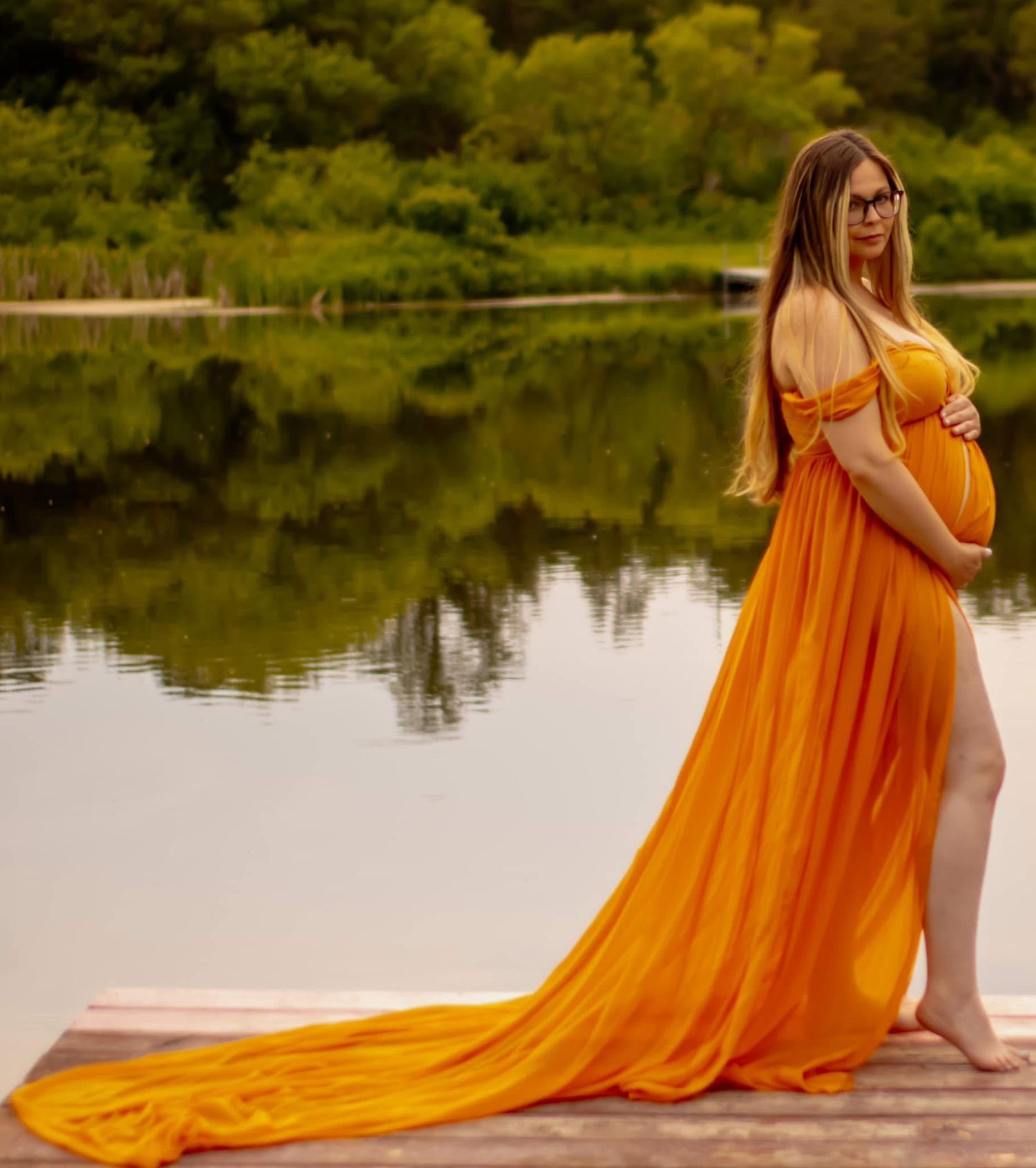 A flowing orange maternity gown with a long train reflects beautifully in a serene pond surrounded by greenery.