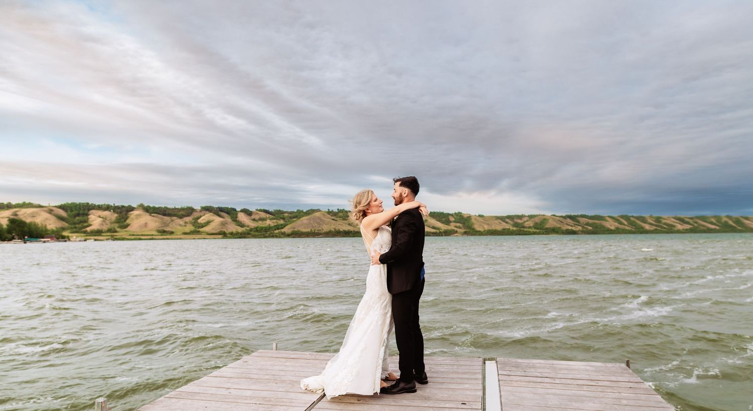 Couple embraces on a wooden dock overlooking a serene lake during sunset.