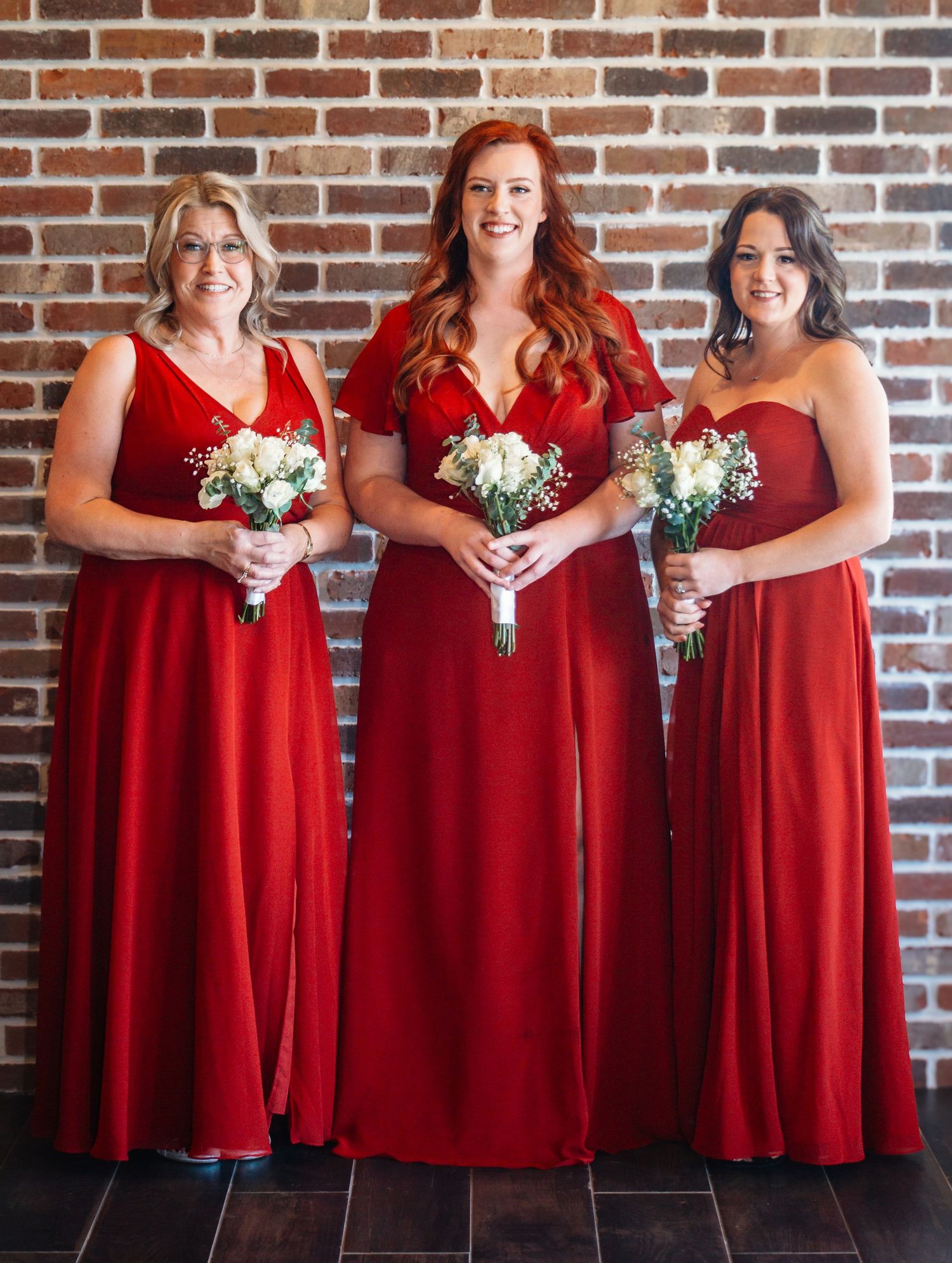 Bridesmaids in matching red dresses stand against a brick wall holding white bouquets.