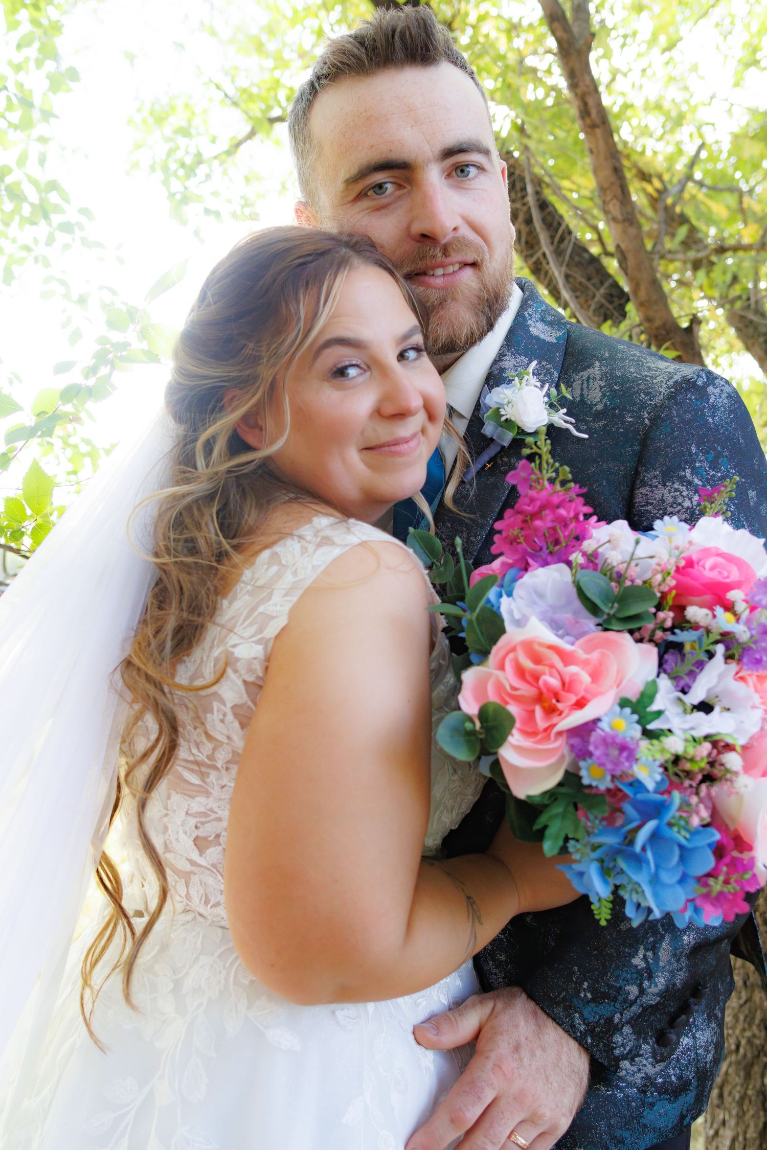 Wedding couple poses with vibrant multicolored bouquet featuring pink and blue flowers.