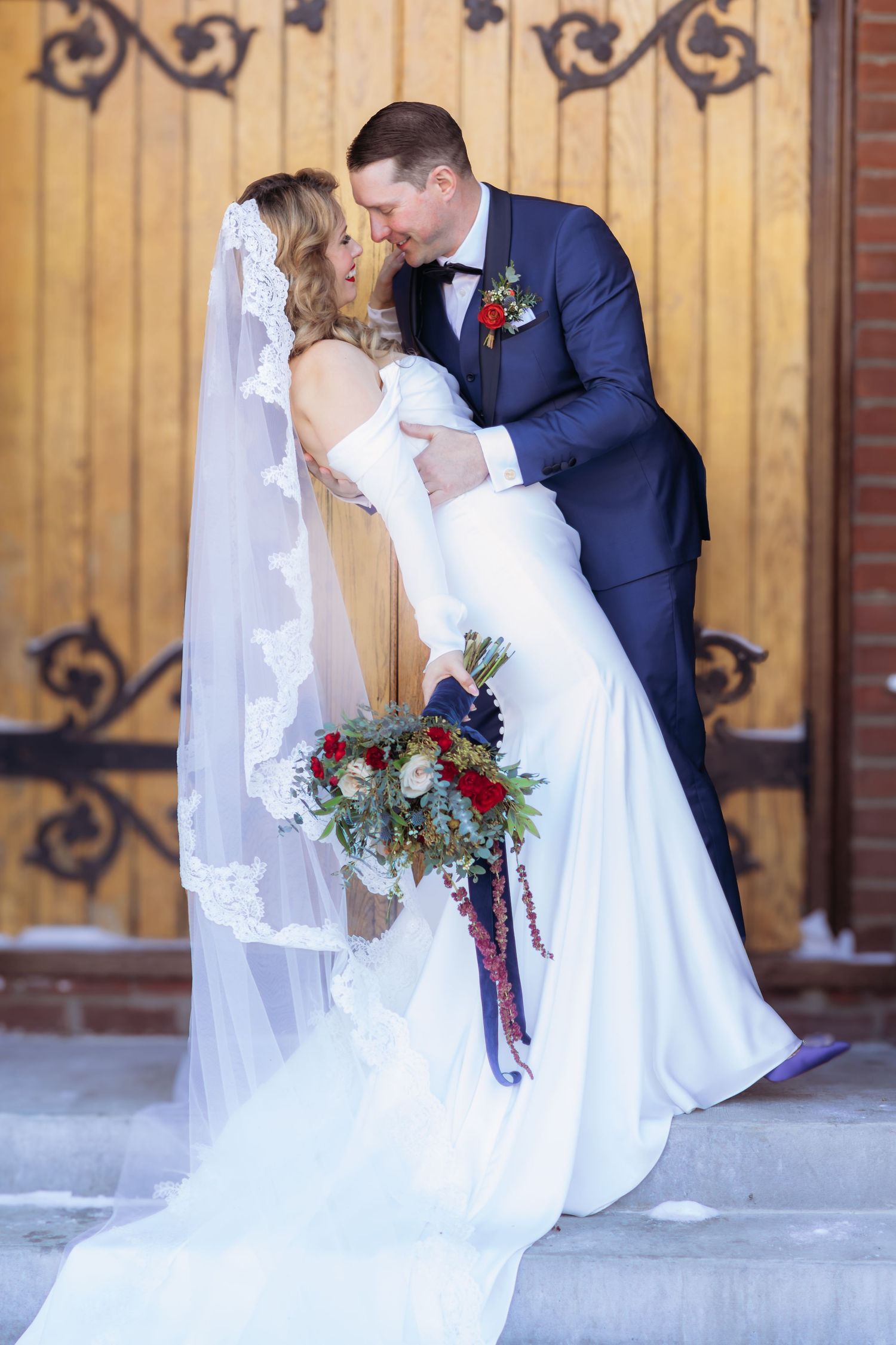 Wedding portrait with flowing veil and red bouquet against ornate wooden doors.