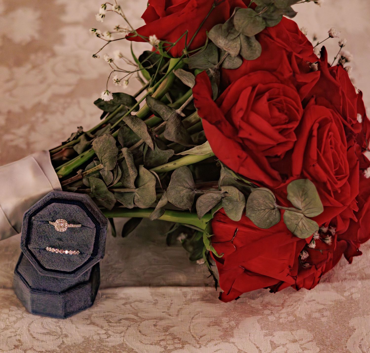 Red roses and wedding rings displayed in navy blue ring boxes on lace fabric.