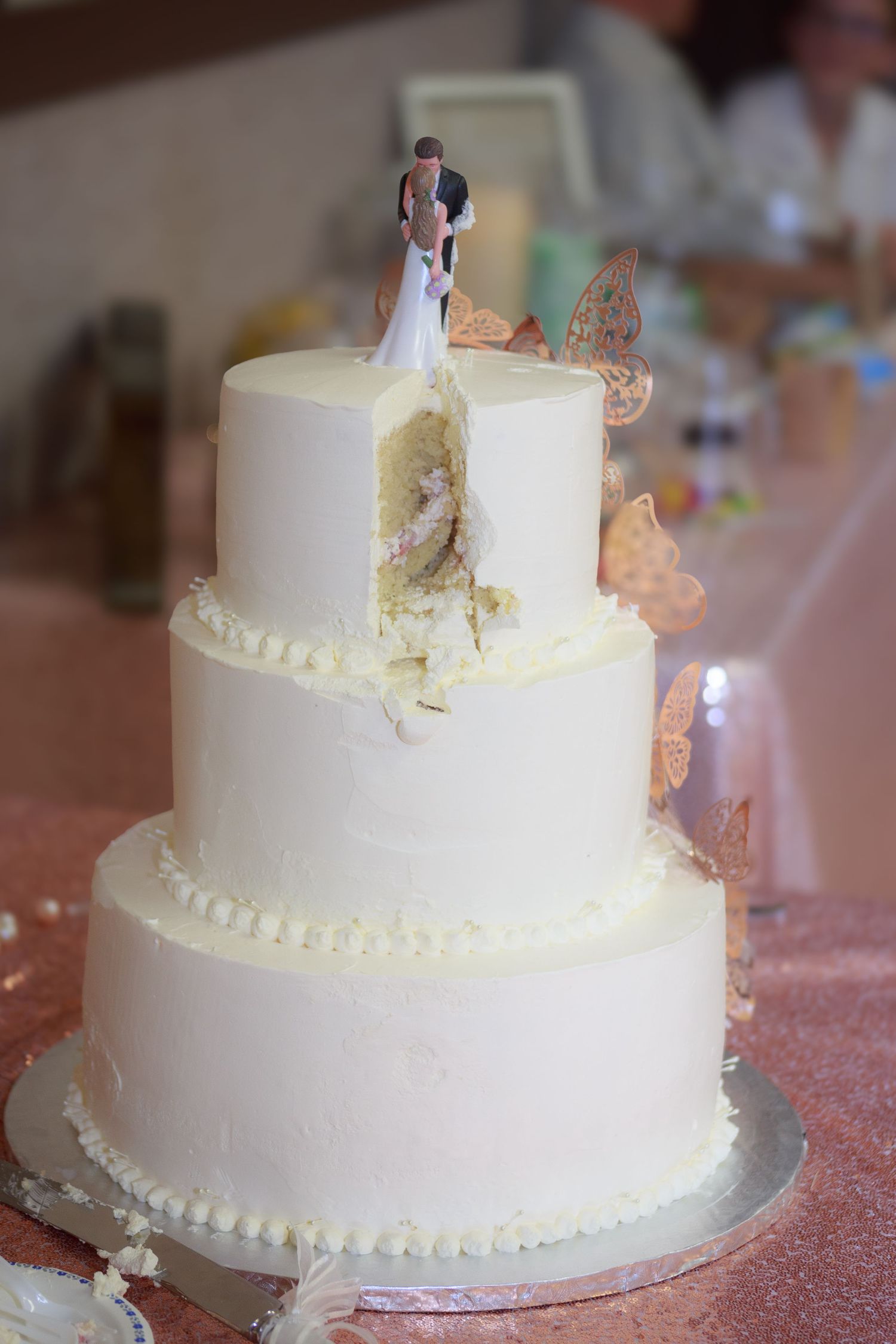 White tiered wedding cake with delicate decorations and miniature topper on sparkly pink tablecloth.
