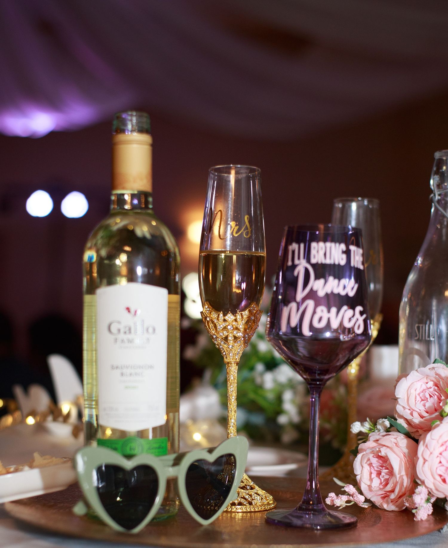 Romantic wine glasses and bottles lit by purple ambient lighting with pink roses.
