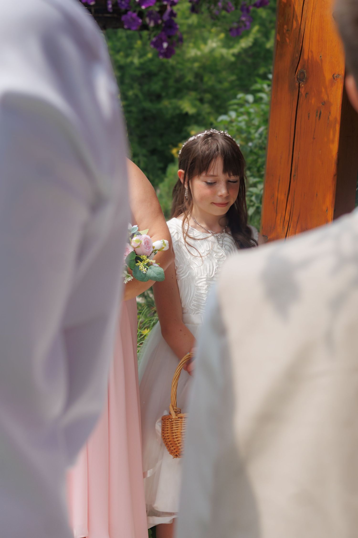 A young attendant in a white dress holds a decorative cone at an outdoor wedding ceremony.