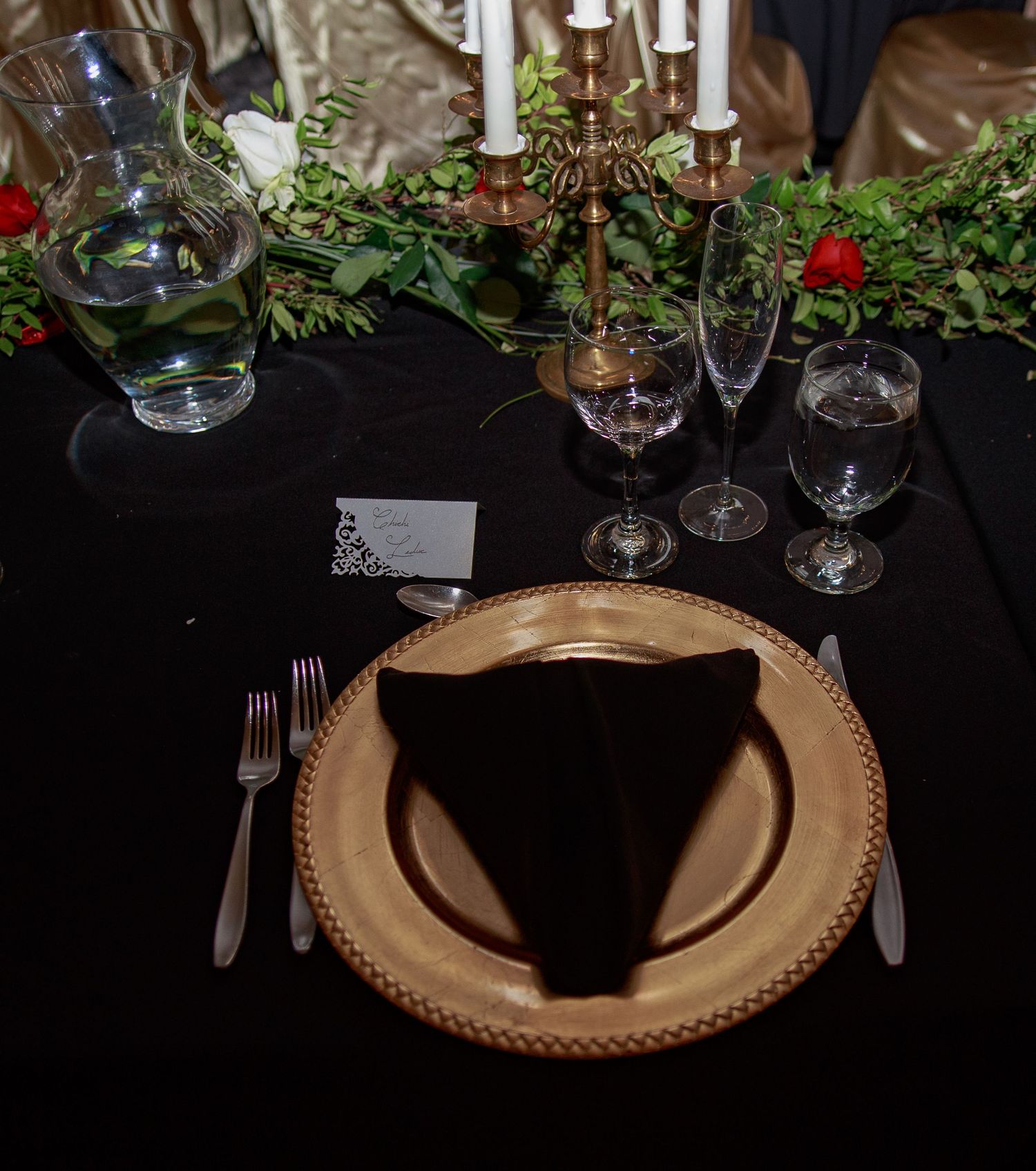 Formal place setting with gold charger plate, black napkin, and greenery centerpiece.