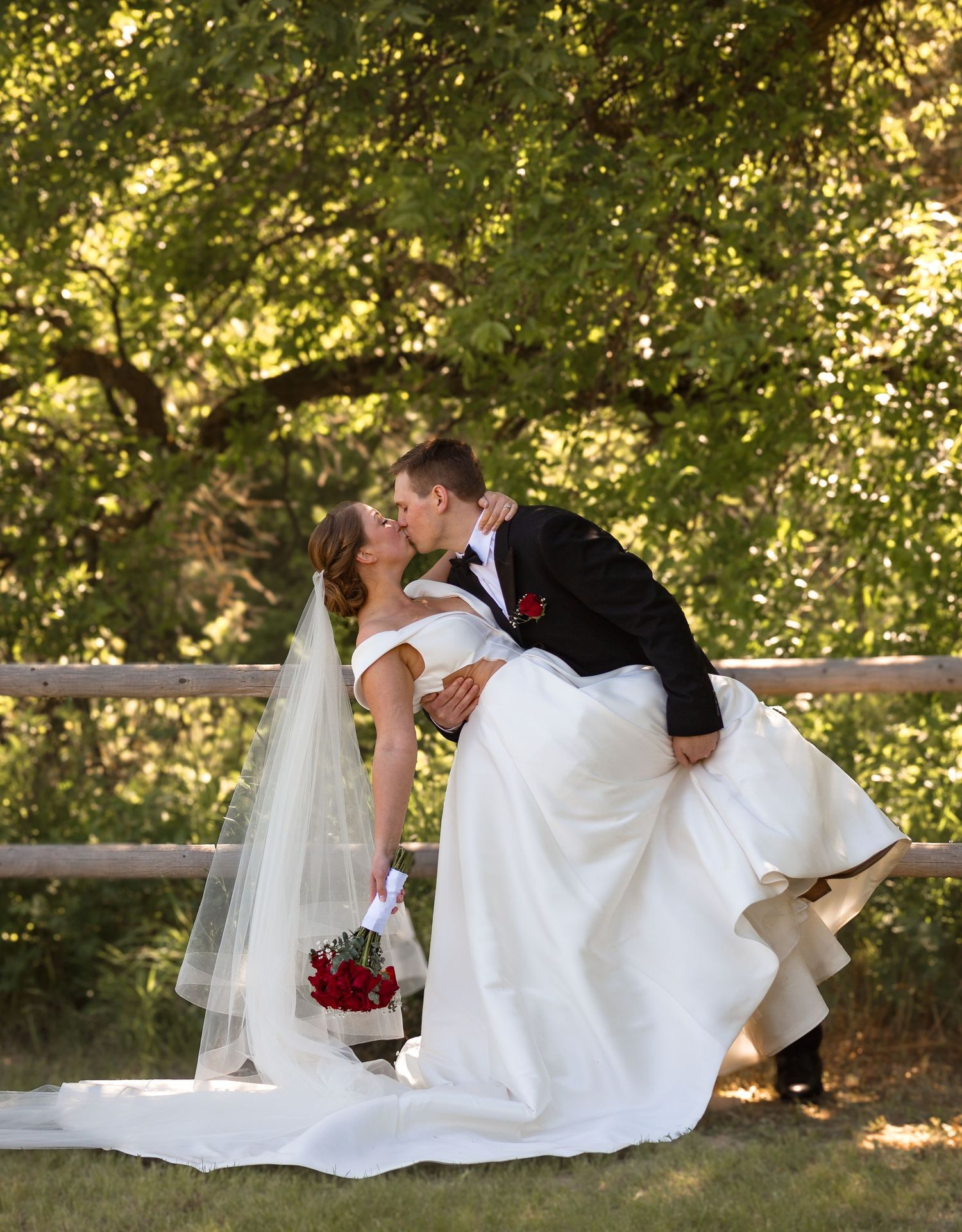 A romantic dip kiss between newlyweds in formal wedding attire against a sunlit garden backdrop.
