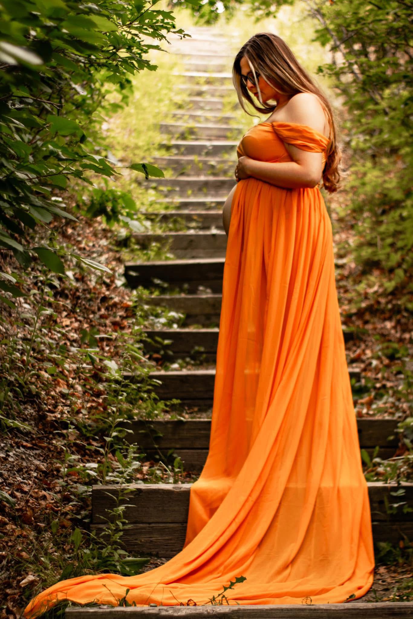 A flowing orange maternity gown cascades down rustic outdoor stairs surrounded by lush greenery.