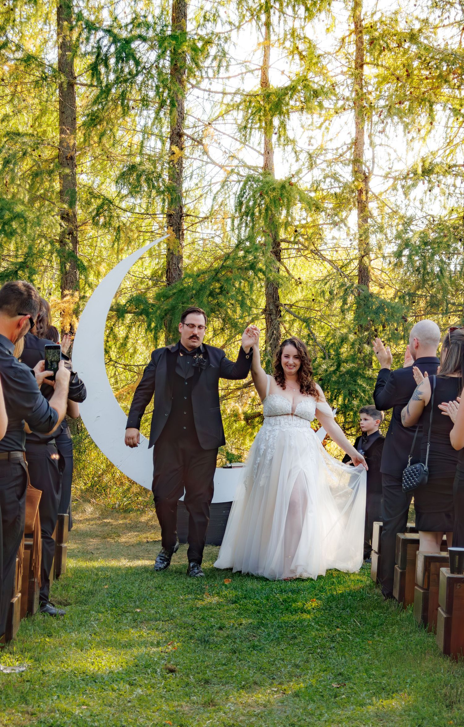 Joyful wedding celebration in a forest setting with guests standing along wooden chairs.