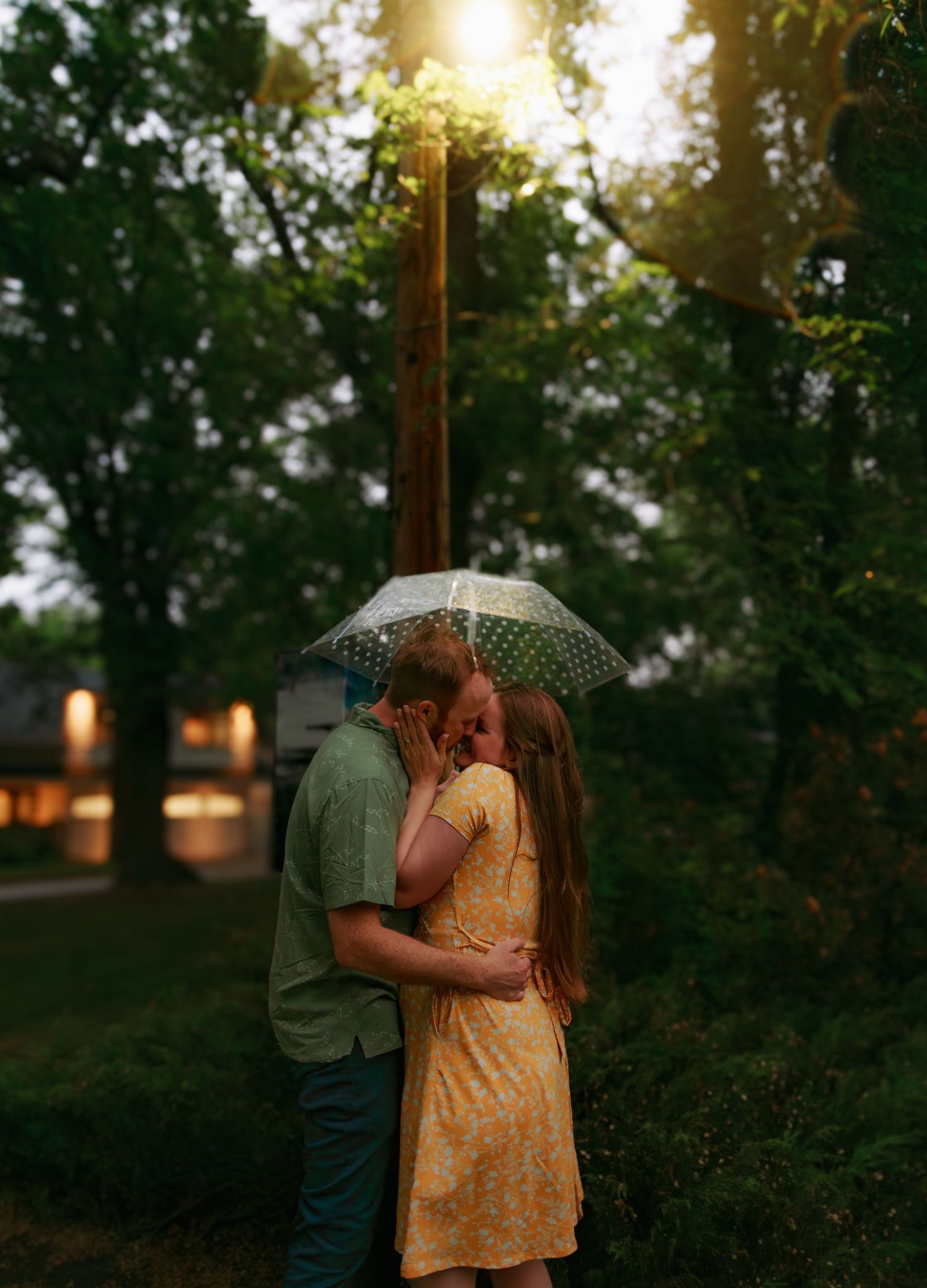 A romantic couple shares an intimate moment under an umbrella at sunset in a wooded area.