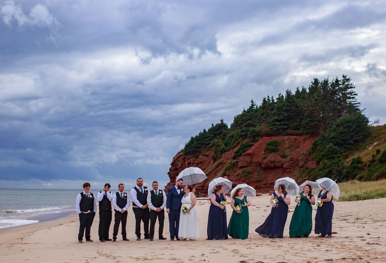 Wedding party poses on a scenic beach with dramatic red cliffs and cloudy skies.