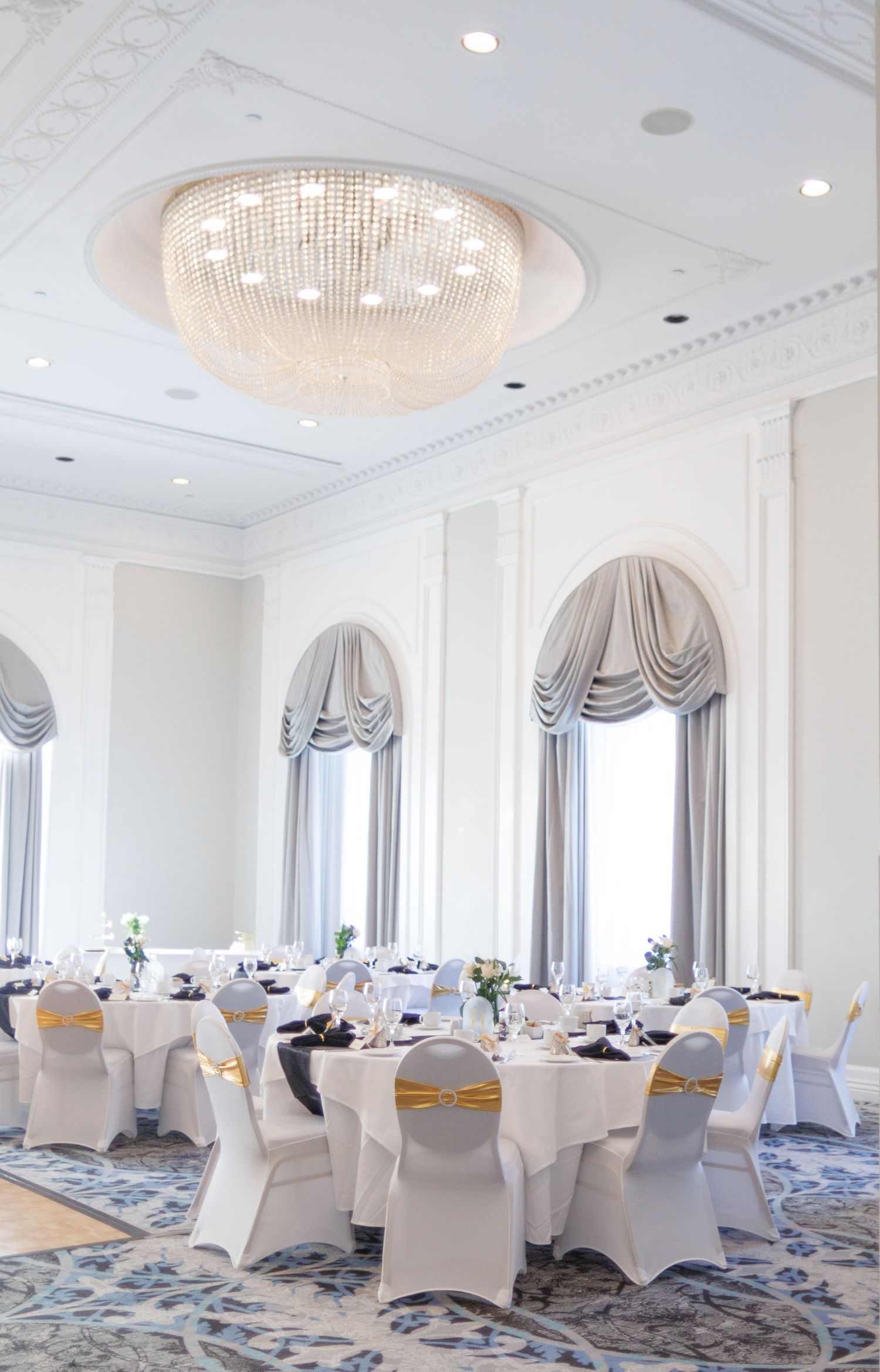 Elegant white ballroom with crystal chandeliers and draped windows set for a wedding reception.