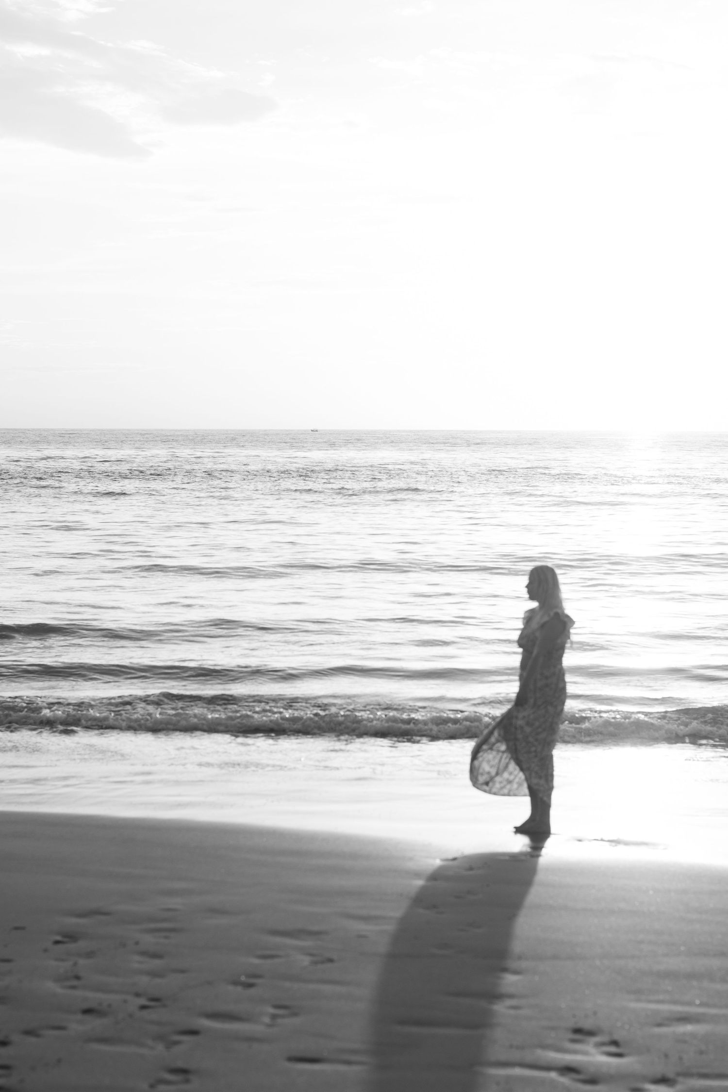 Black and white silhouette of person walking along misty beach at sunset.