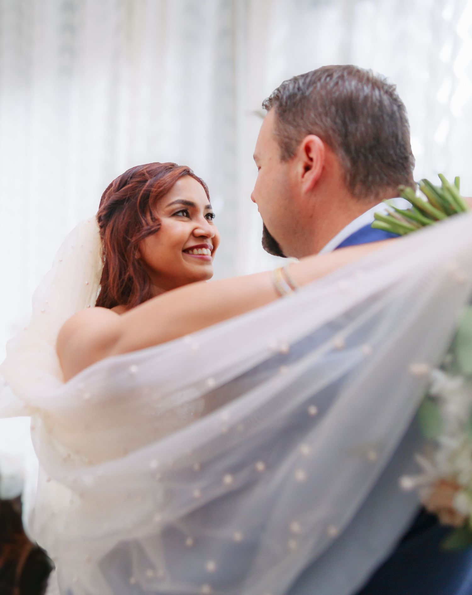 Elegant wedding couple embracing behind flowing white veil in indoor setting.