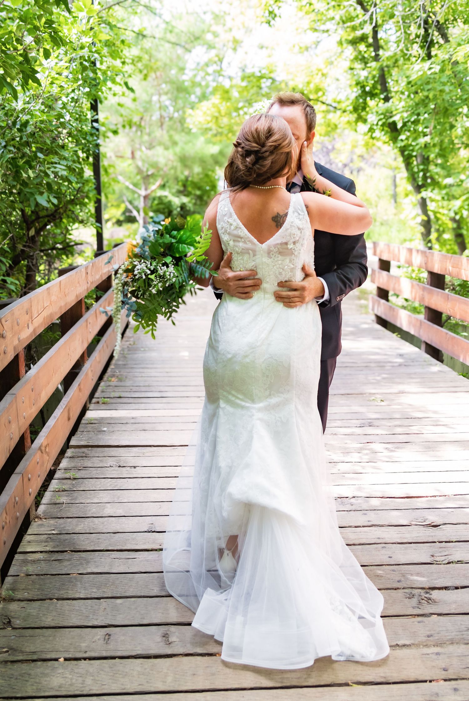 Wedding couple poses on a wooden boardwalk surrounded by lush green trees and natural lighting.