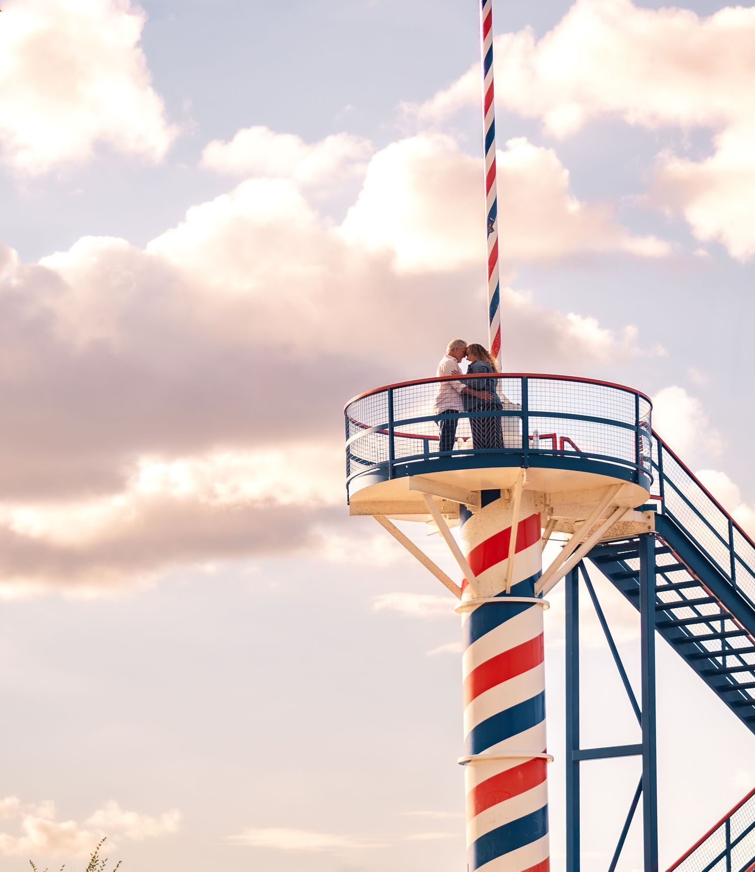 Person standing on red and white striped lighthouse platform against sunset sky.
