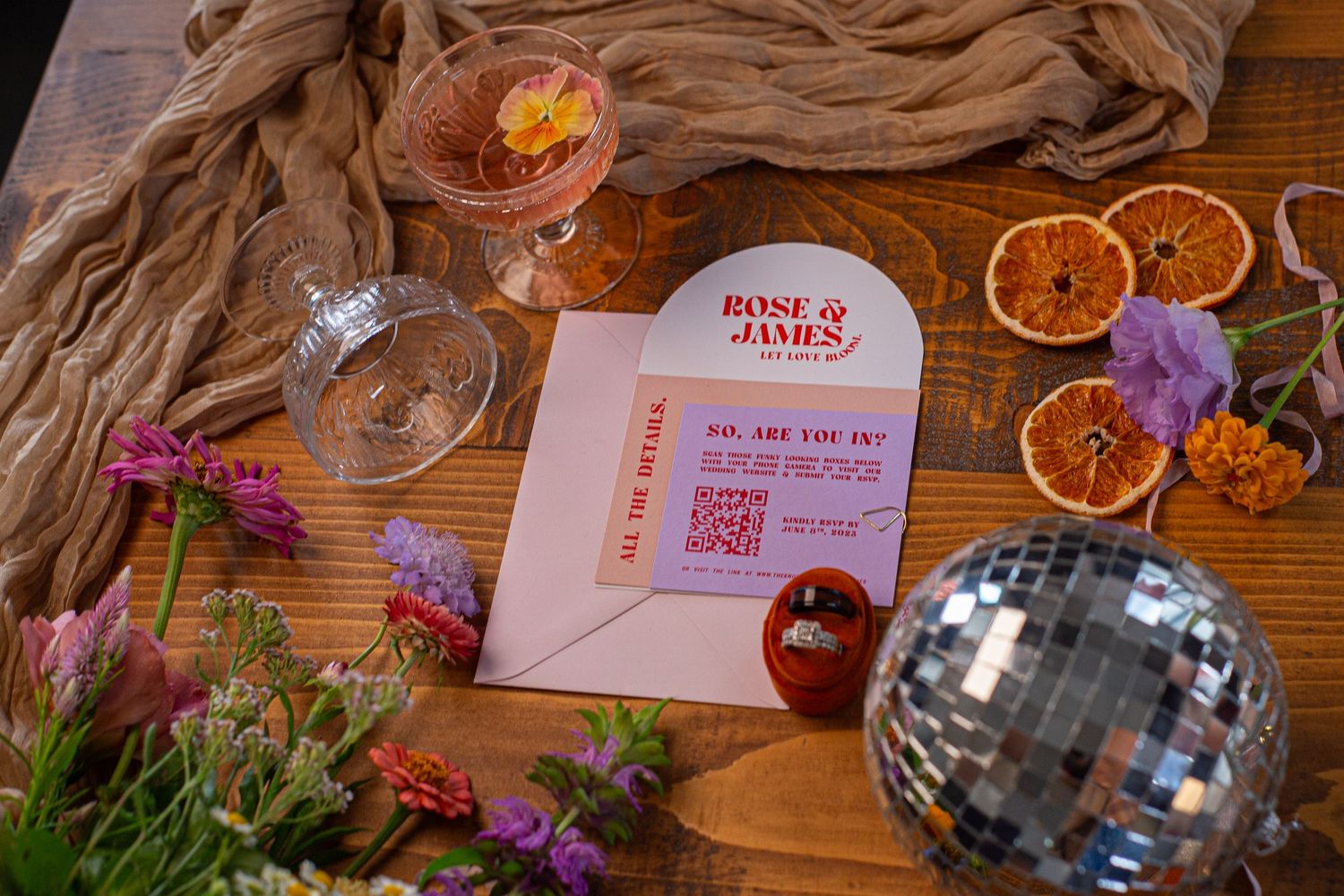 Wedding reception details with disco ball, dried oranges, and pink stationery on rustic wooden table.