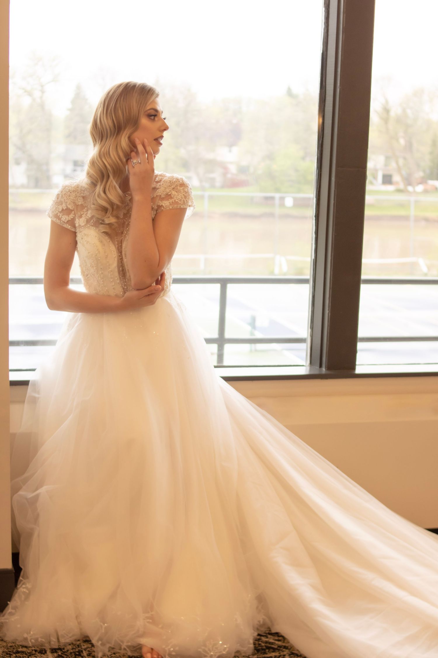 Flowing white wedding gown with delicate details photographed by window in natural light.