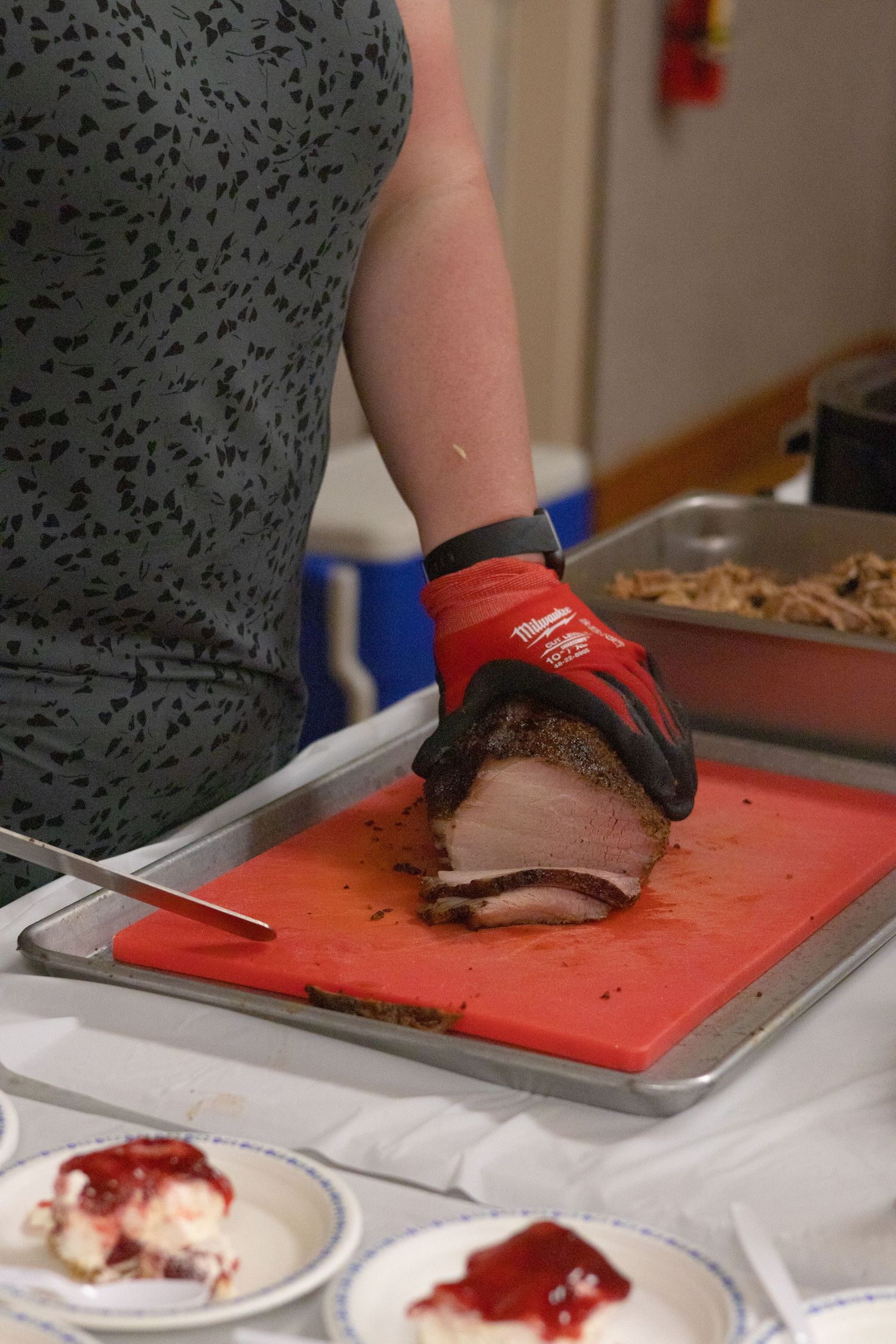Hands in black gloves slice meat on red cutting board at catering station.