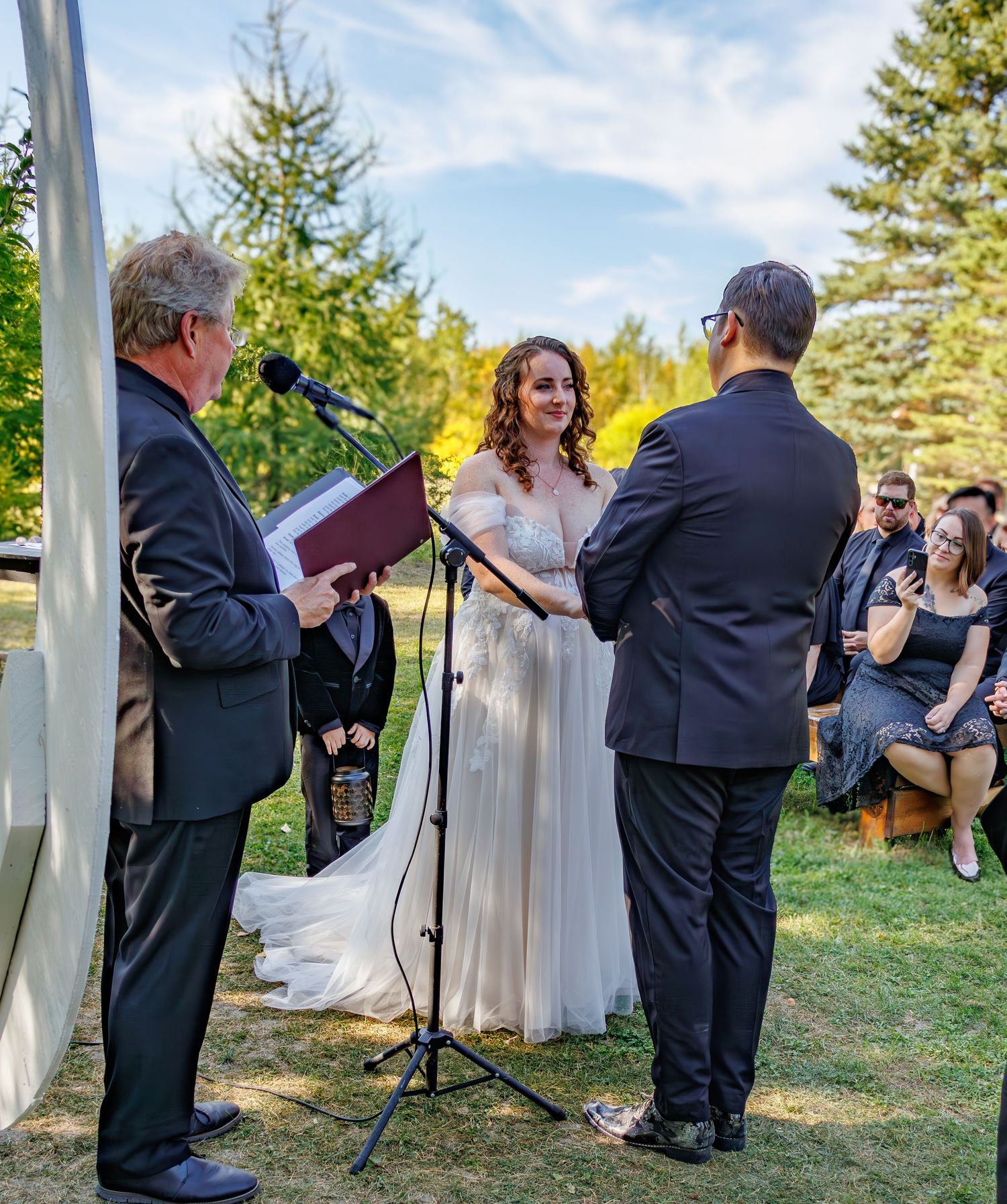 Outdoor wedding ceremony with guests seated on wooden chairs among trees.