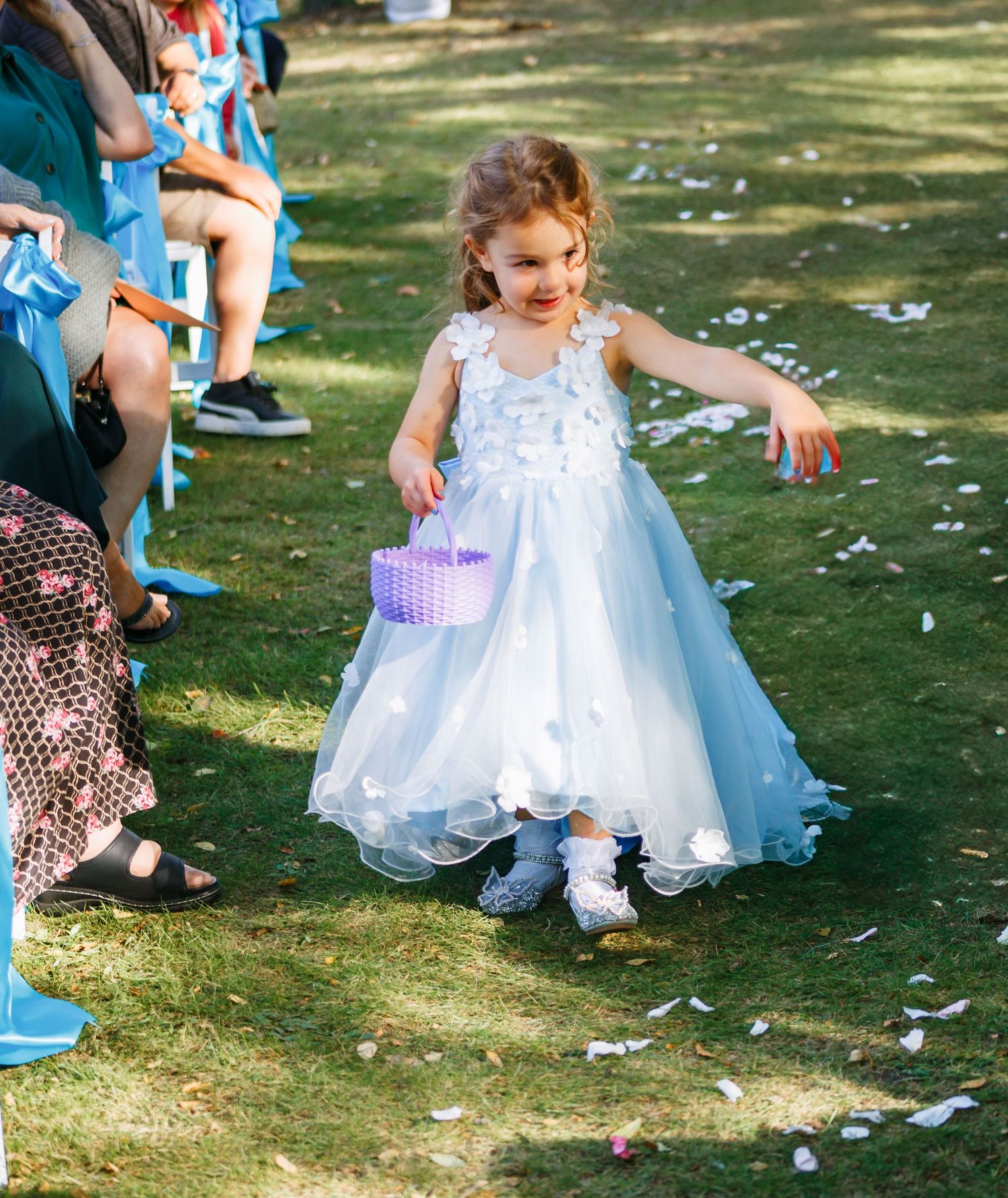 Flower girl in white tulle dress plays with petals on grass during outdoor wedding celebration.