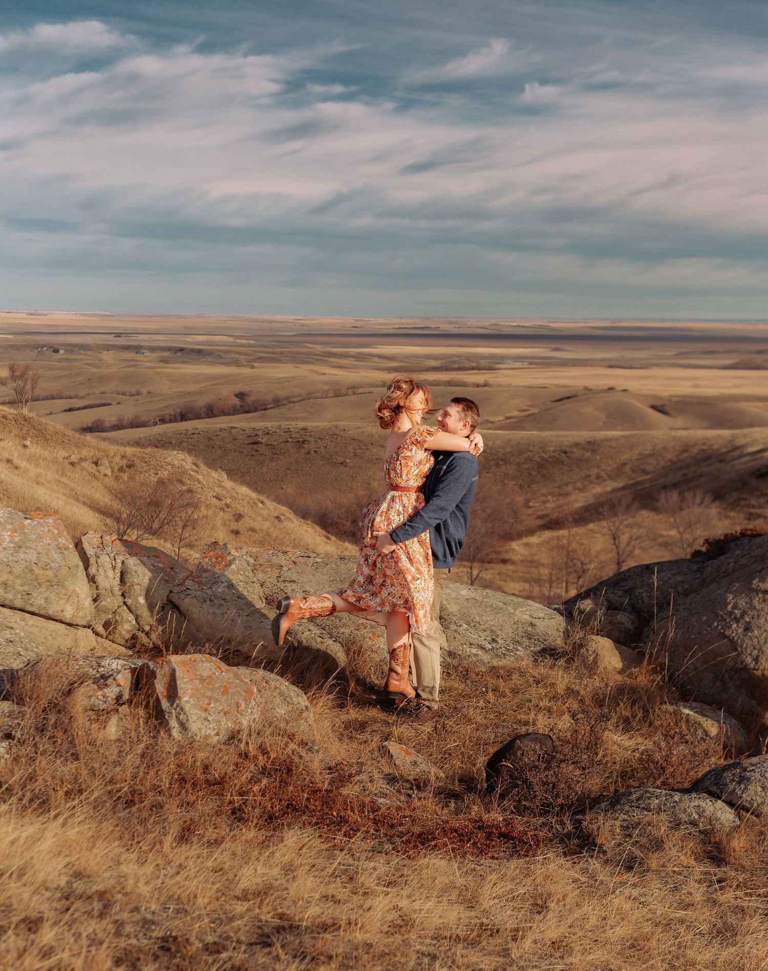 Romantic embrace on a rocky cliff overlooking vast rolling hills during sunset.