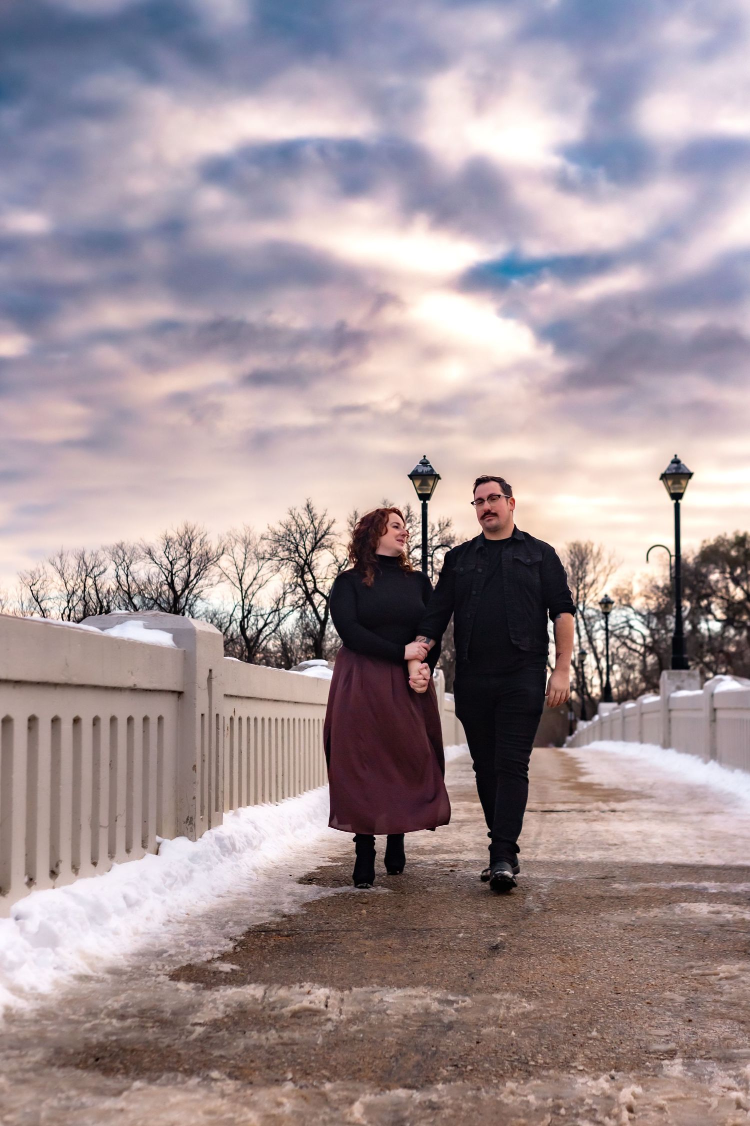 Couple walking along snow-covered path with white fence and lamp posts at dusk.