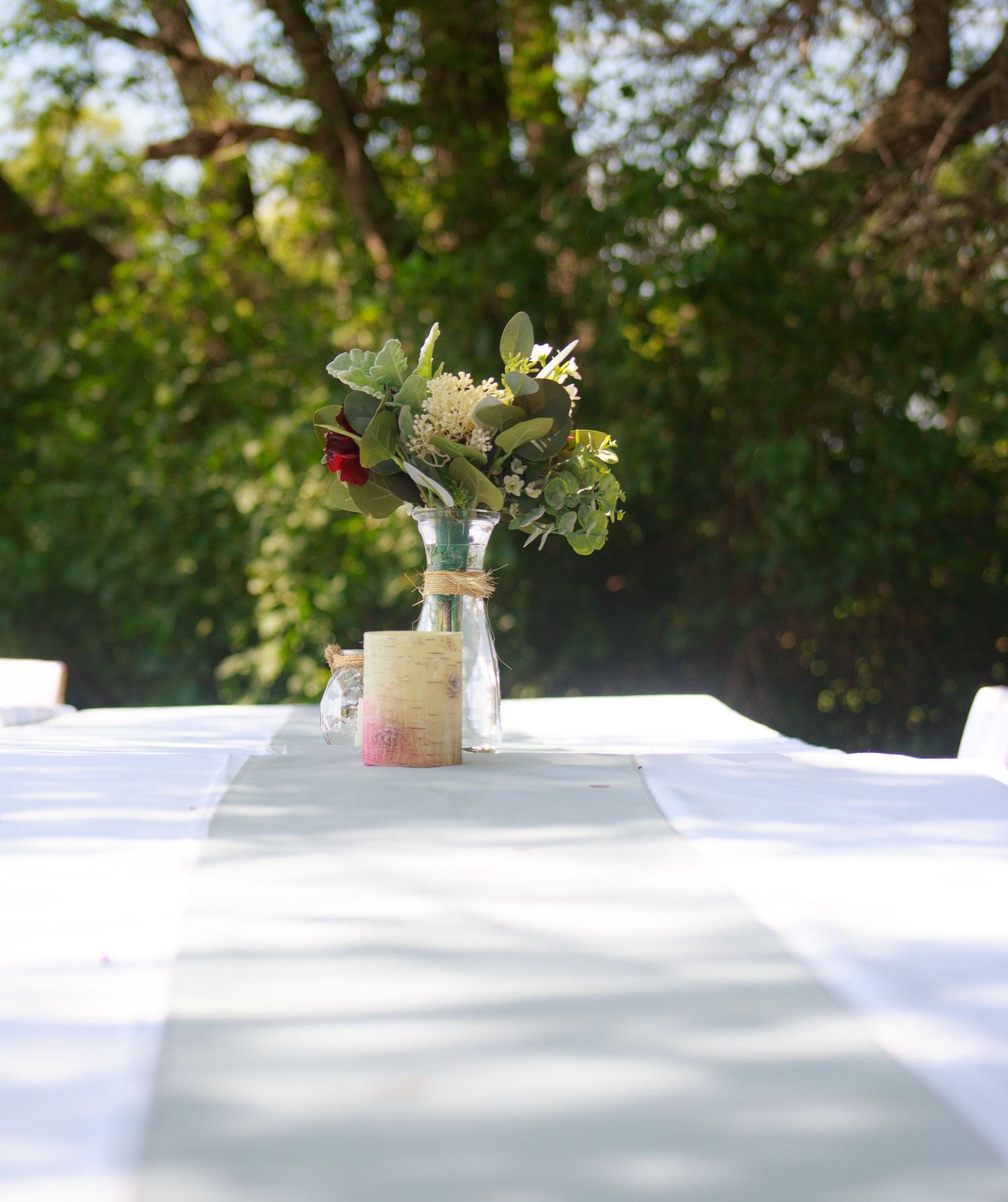 Clean white tablecloth with simple floral centerpiece outdoors under trees.