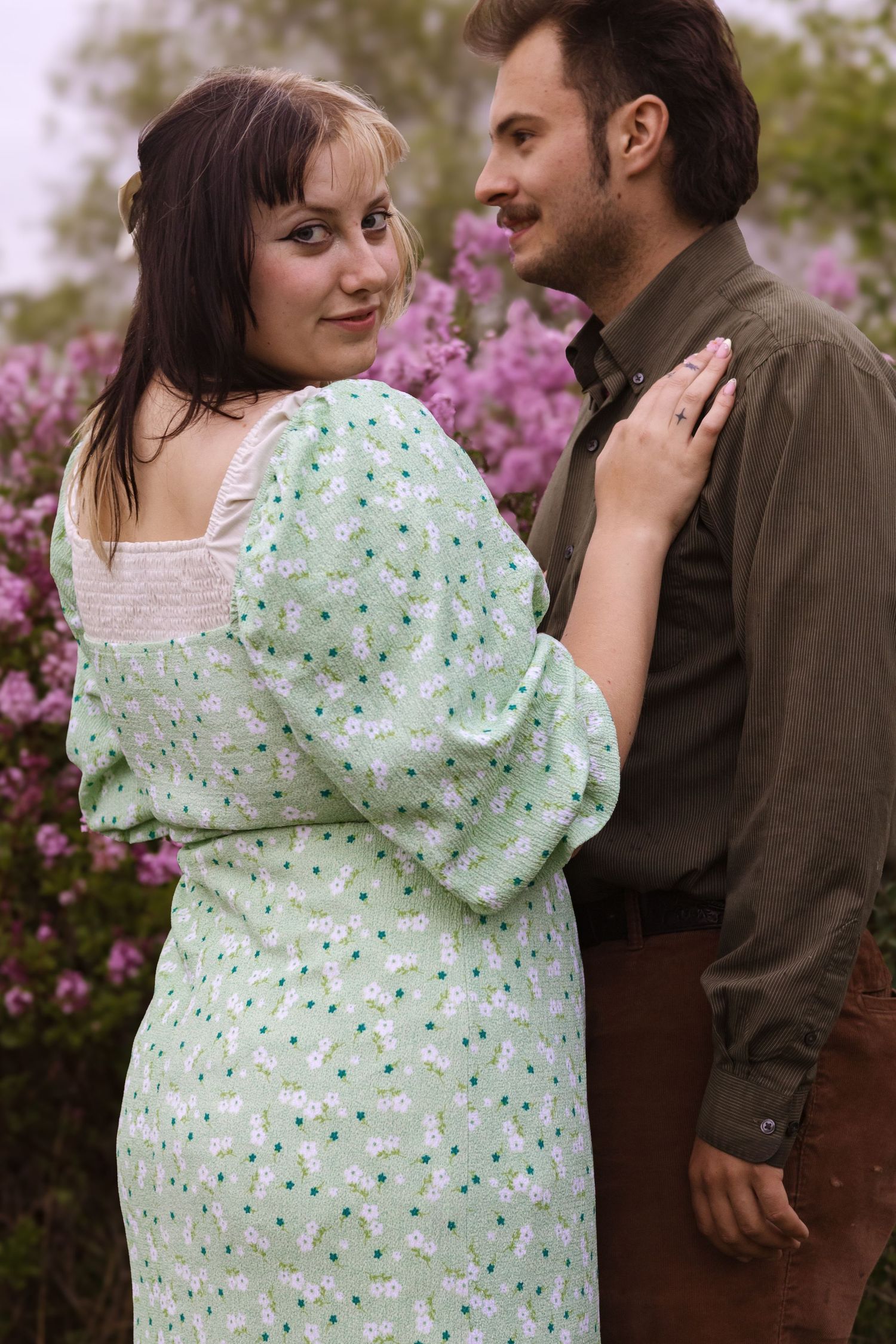A couple in a mint floral dress and brown shirt share tender moments near blooming purple flowers.