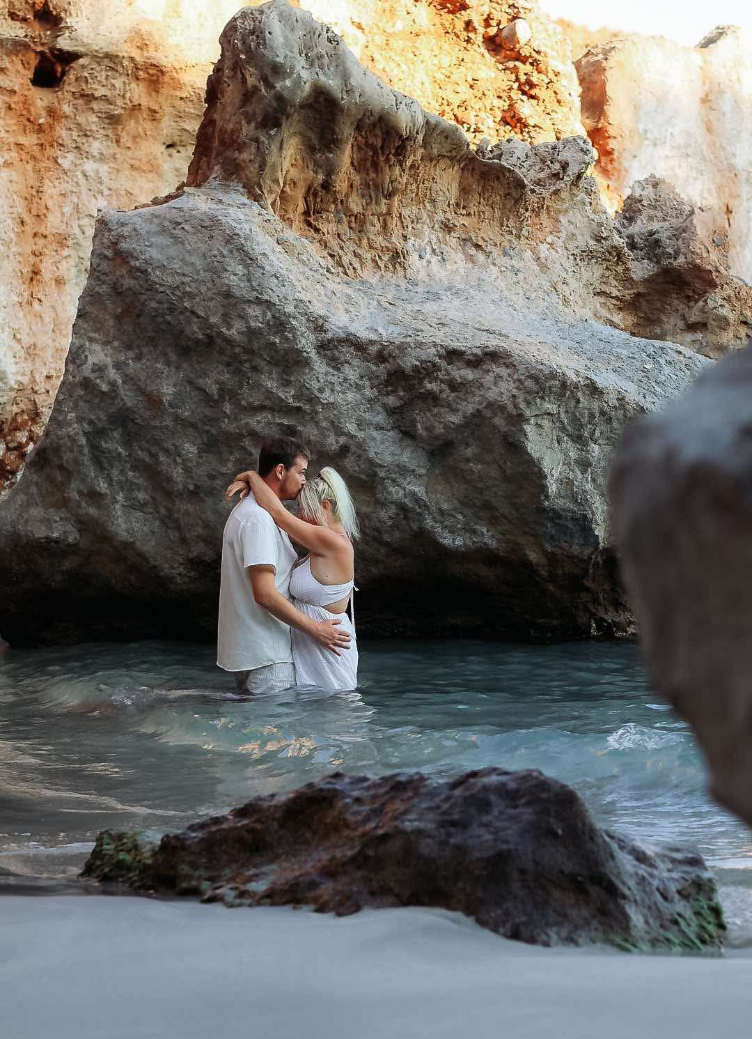 Couple embraces romantically on a rocky beach near ocean caves at sunset.