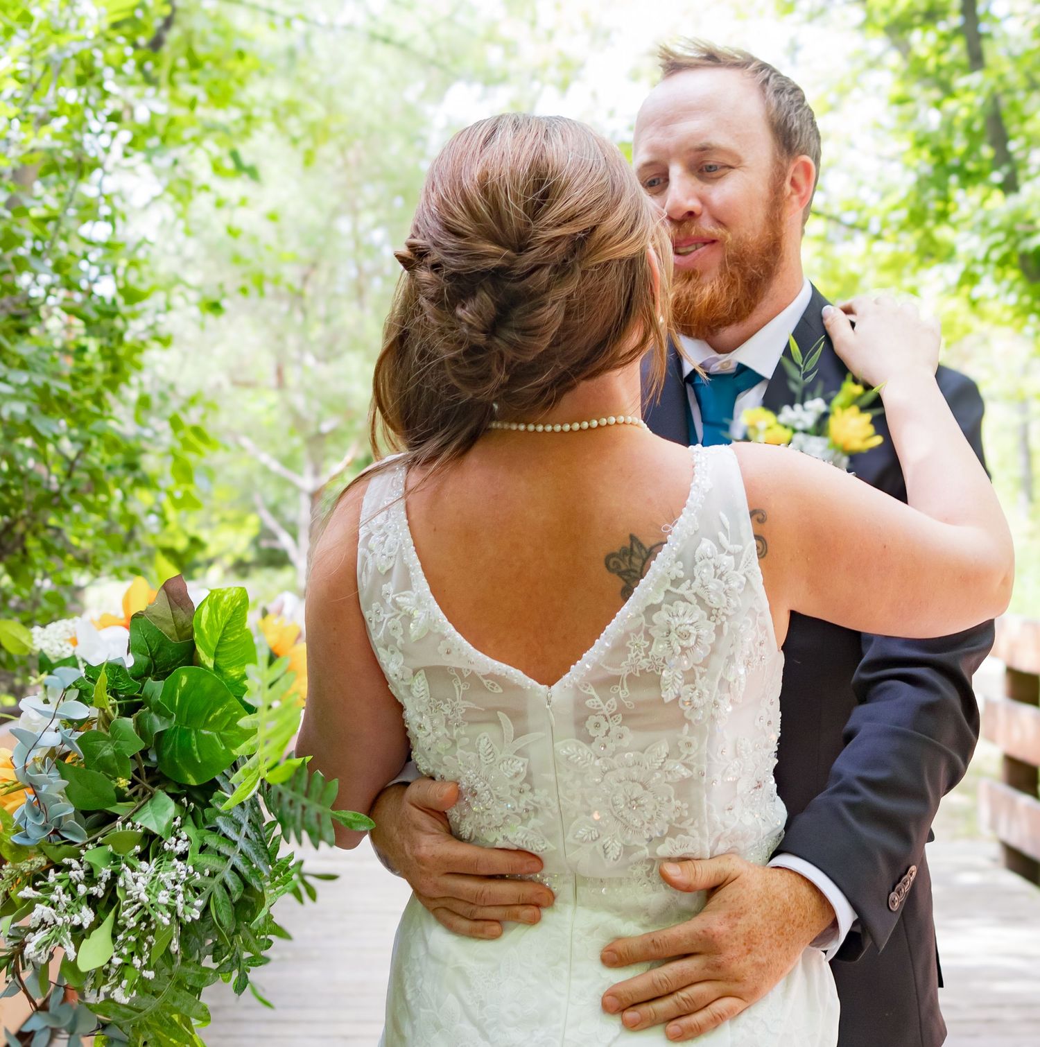 Romantic wedding couple embraces on a wooden bridge pathway surrounded by greenery.