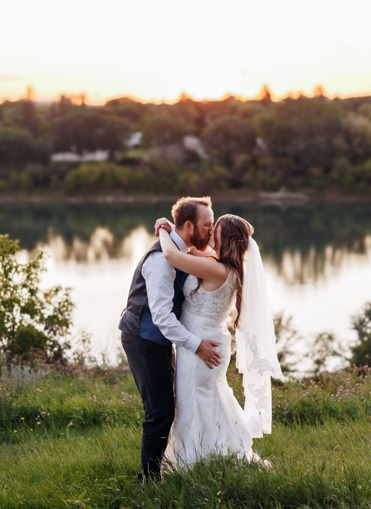 Wedding couple embracing by a serene lake during sunset.