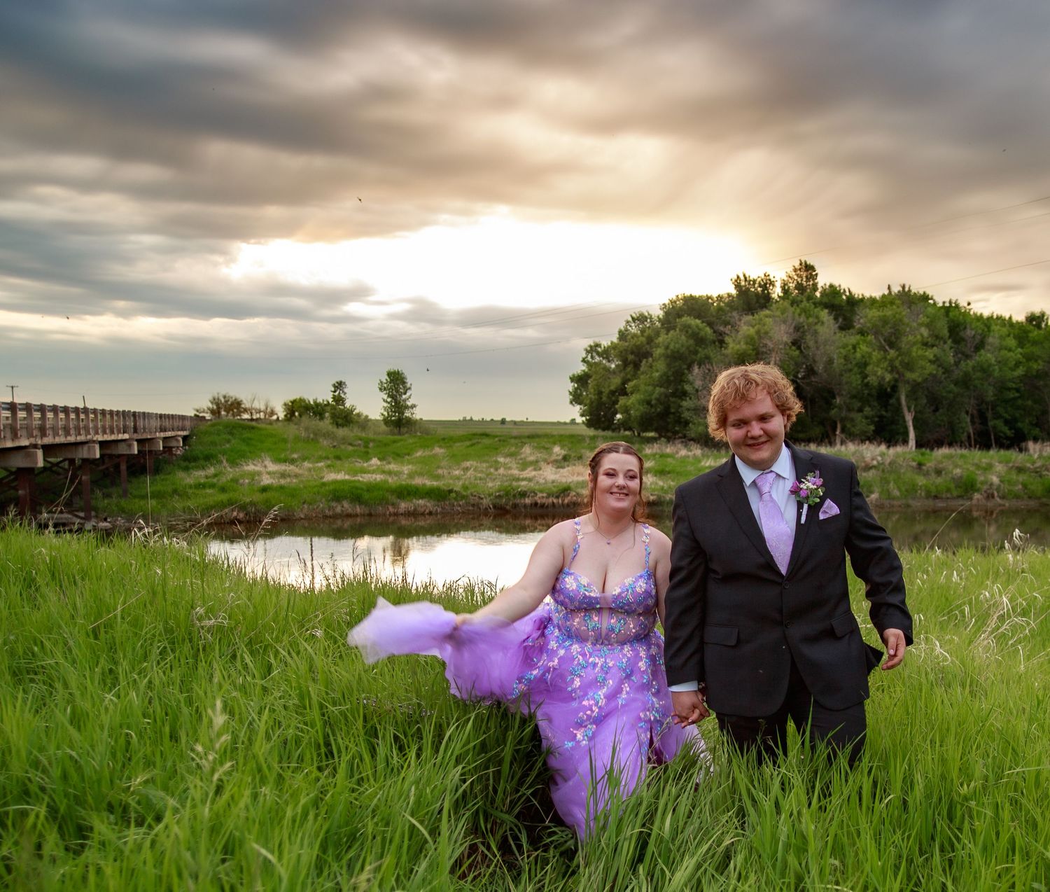 A couple poses together in formal attire against a scenic rural landscape at sunset.