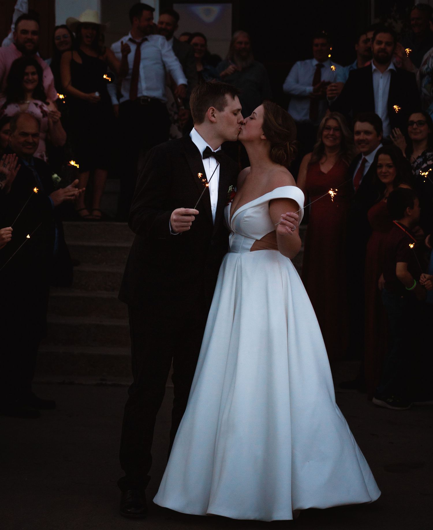 A romantic evening wedding kiss surrounded by guests holding sparklers.