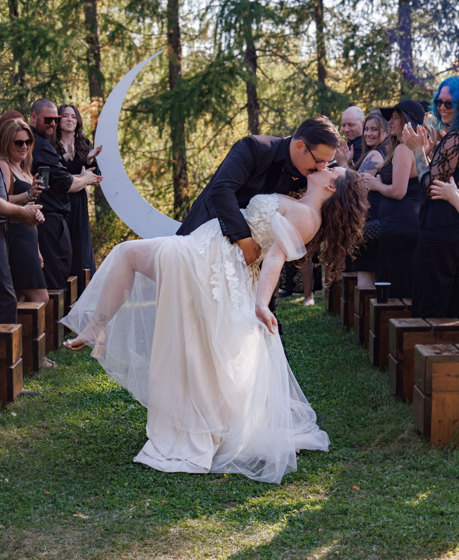 Wedding ceremony guests seated on wooden benches under tall evergreen trees.
