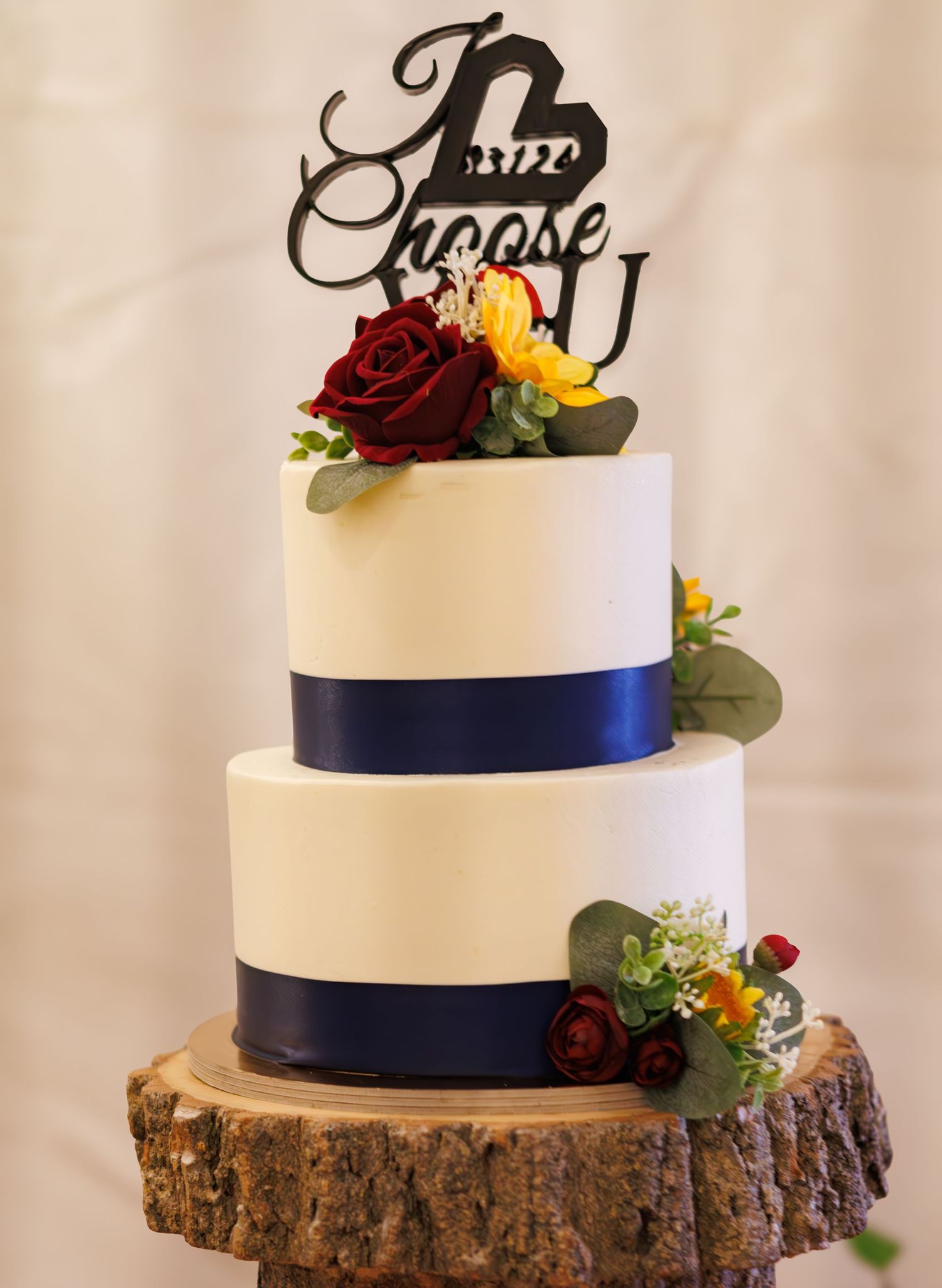 Two-tier white wedding cake with navy ribbon and floral decorations on wood slice base.
