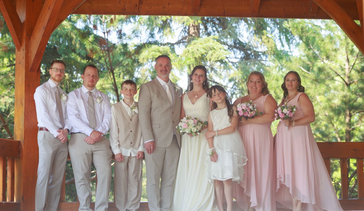 Wedding party in pink and beige attire poses under a wooden pavilion for formal photos.