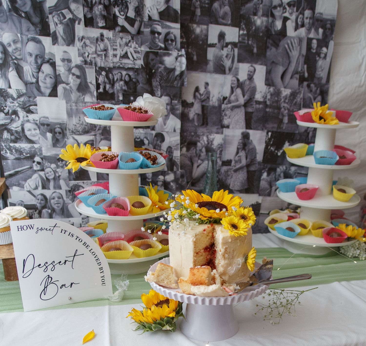 Dessert bar with colorful cupcakes and sunflower decorated cake against black and white photo wall.