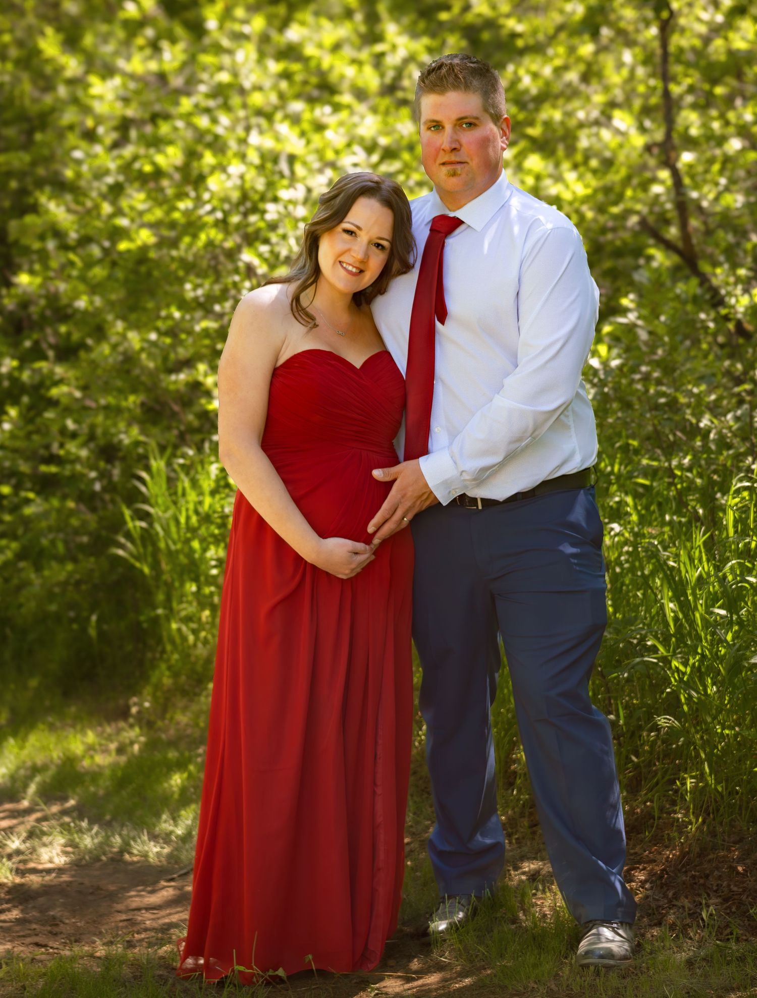 Couple poses intimately in formal wear with red dress against green foliage.