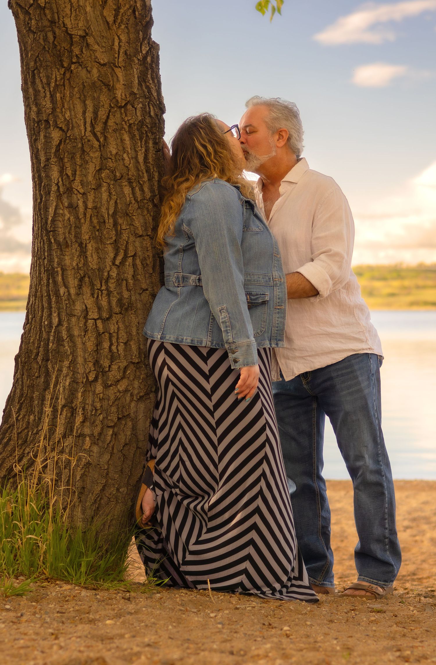 Couple sharing intimate moment against tree trunk with striped dress visible in golden light.