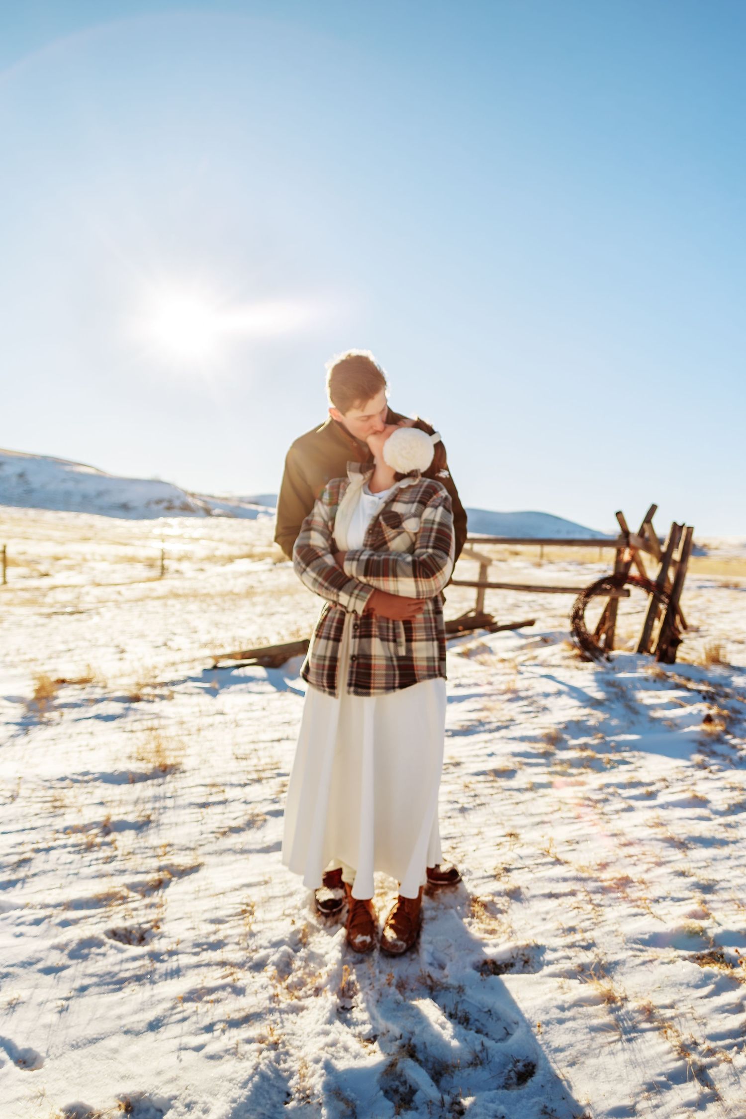 A couple embraces in winter snow with mountains and sunlight in the background.
