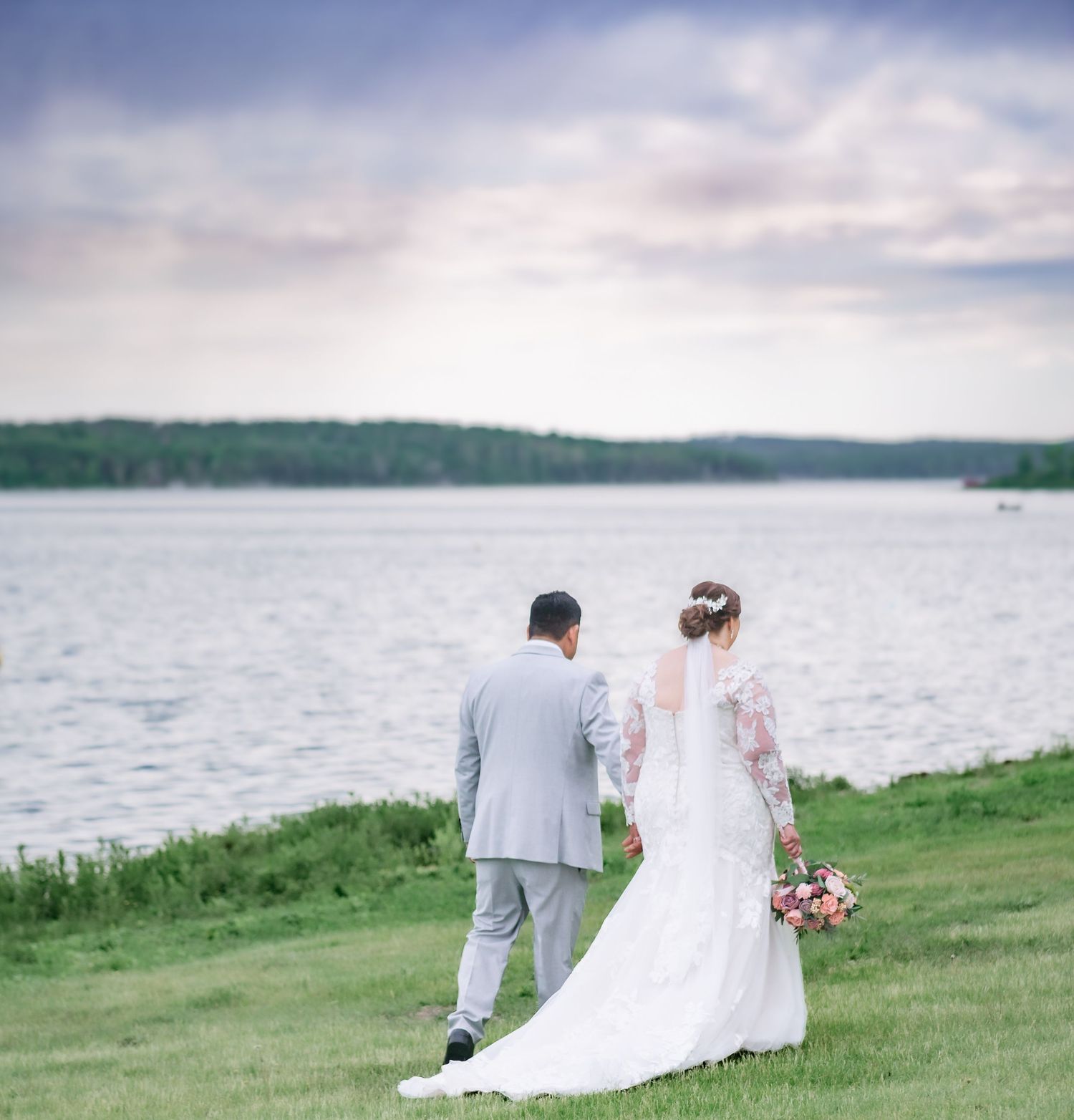 A serene lakeside wedding portrait showing couple walking along grassy shore at sunset.
