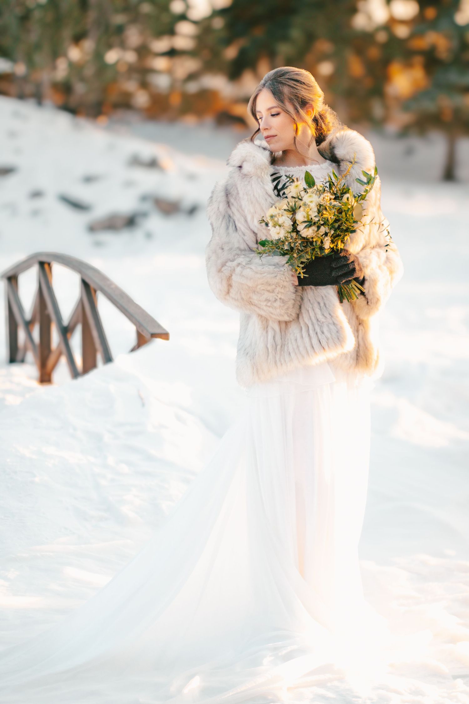 A bride in a white dress holds a bouquet while standing in fresh snow during golden hour.