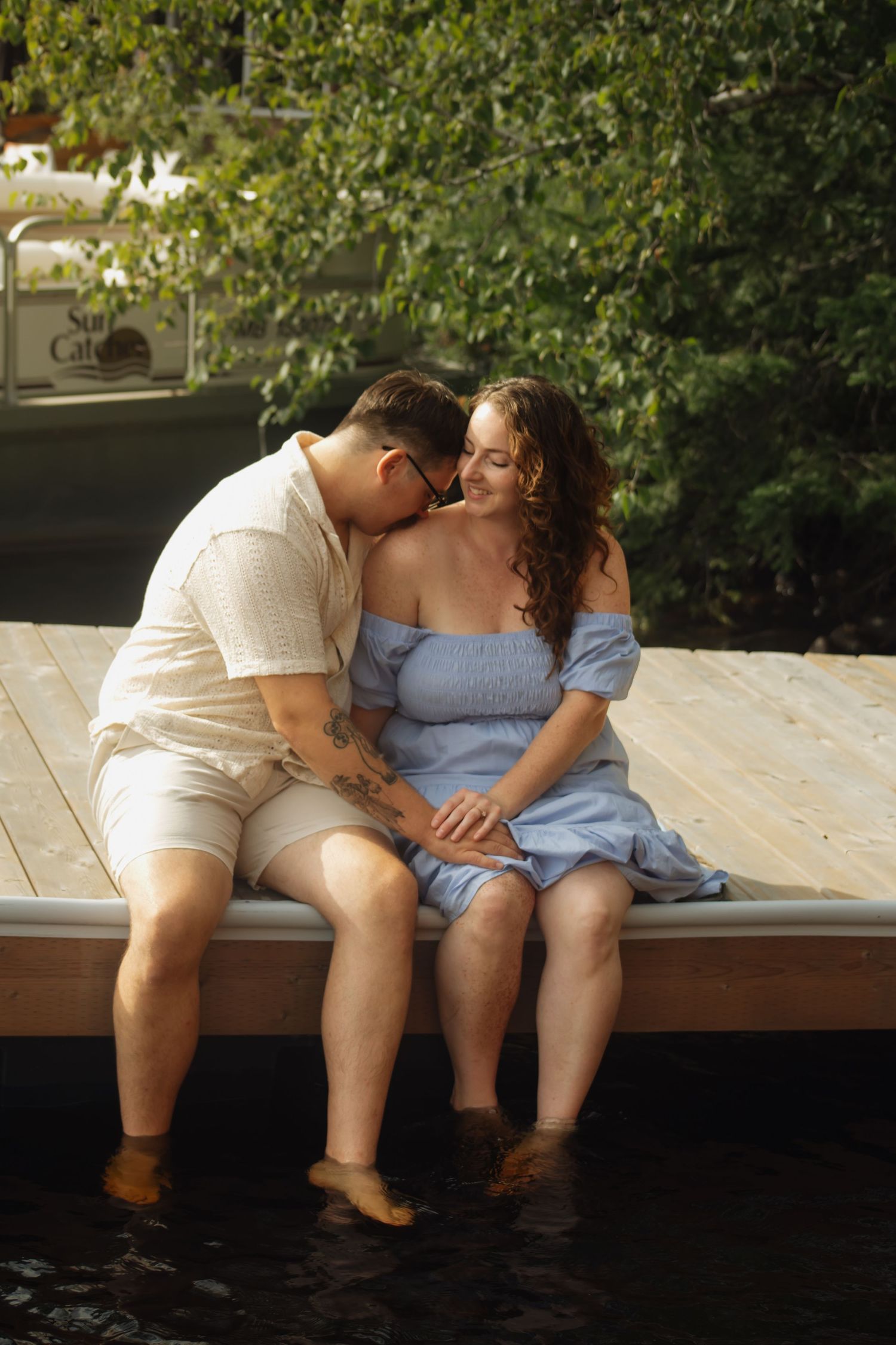 A romantic couple sits together on a wooden dock by the water on a sunny day.