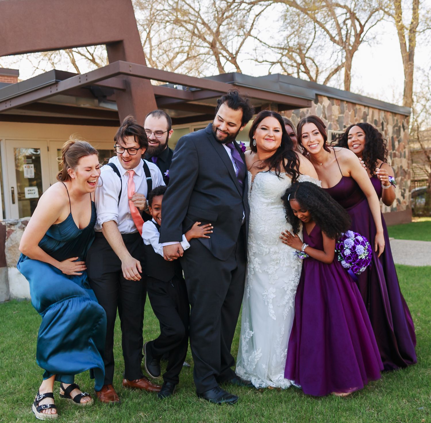 Wedding party strikes fun poses together on grassy lawn wearing purple dresses and black suits.