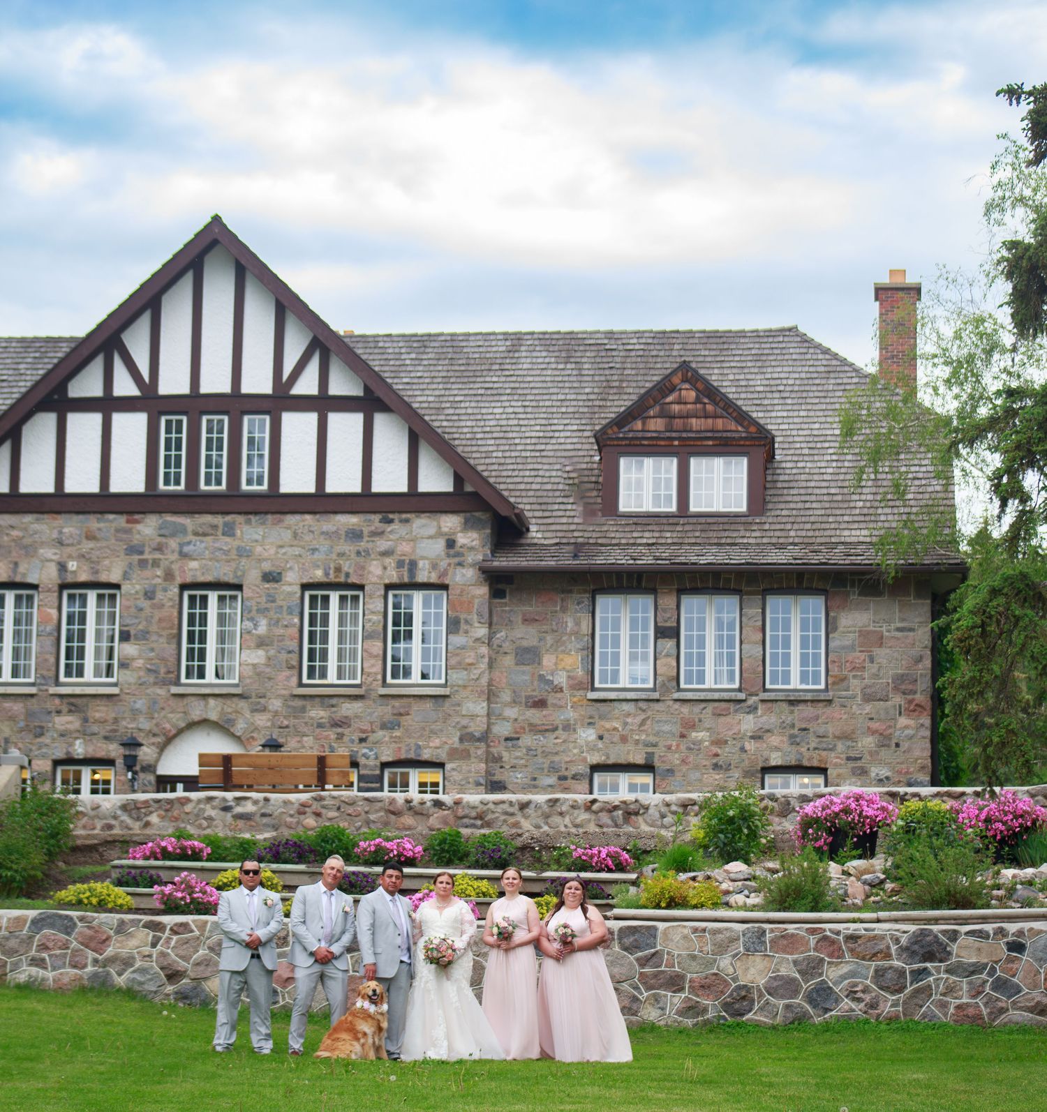 Wedding party stands in front of stone and Tudor-style mansion with pink flowers in bloom.