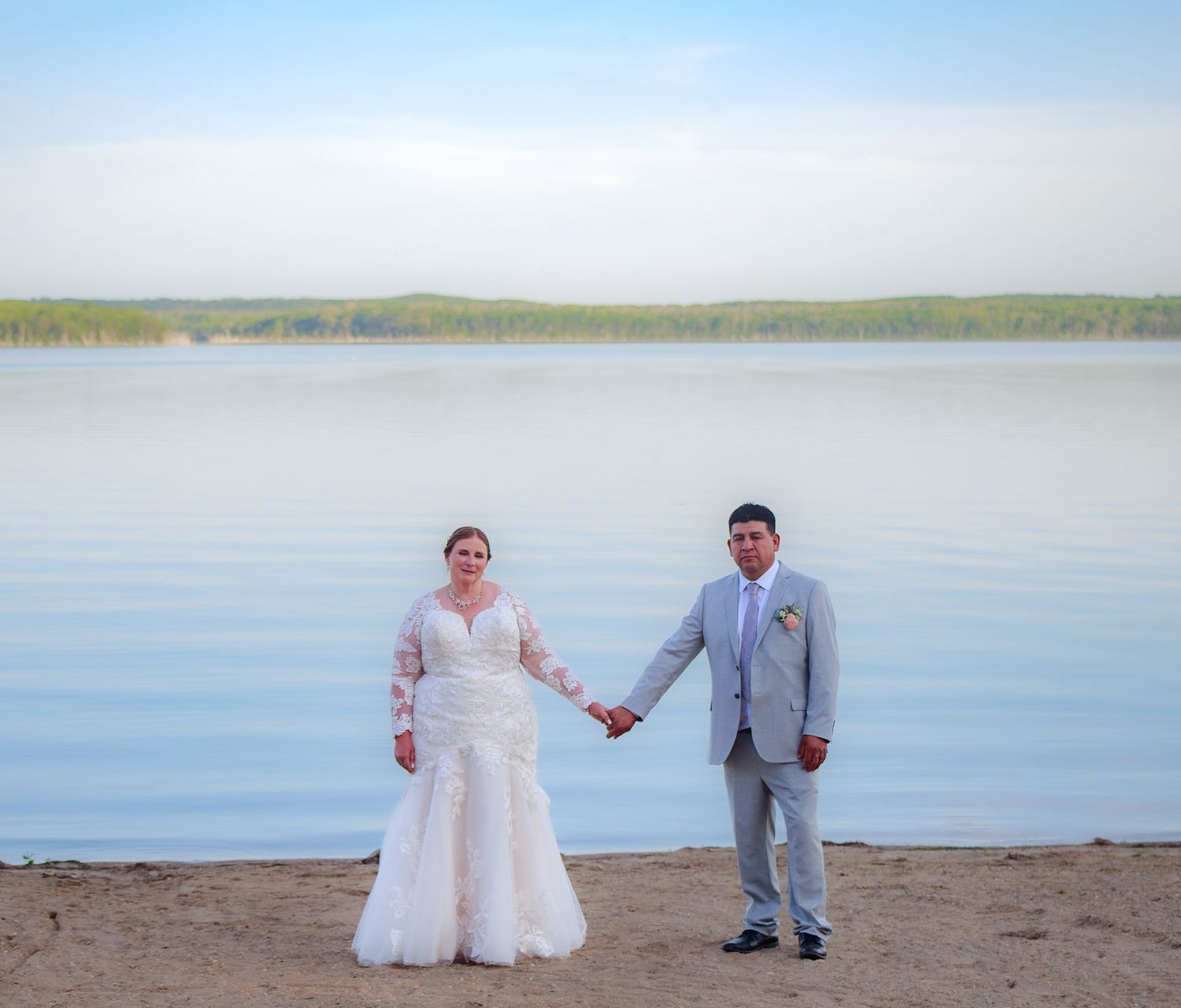 Wedding couple poses together on a sandy beach with calm lake waters in the background.