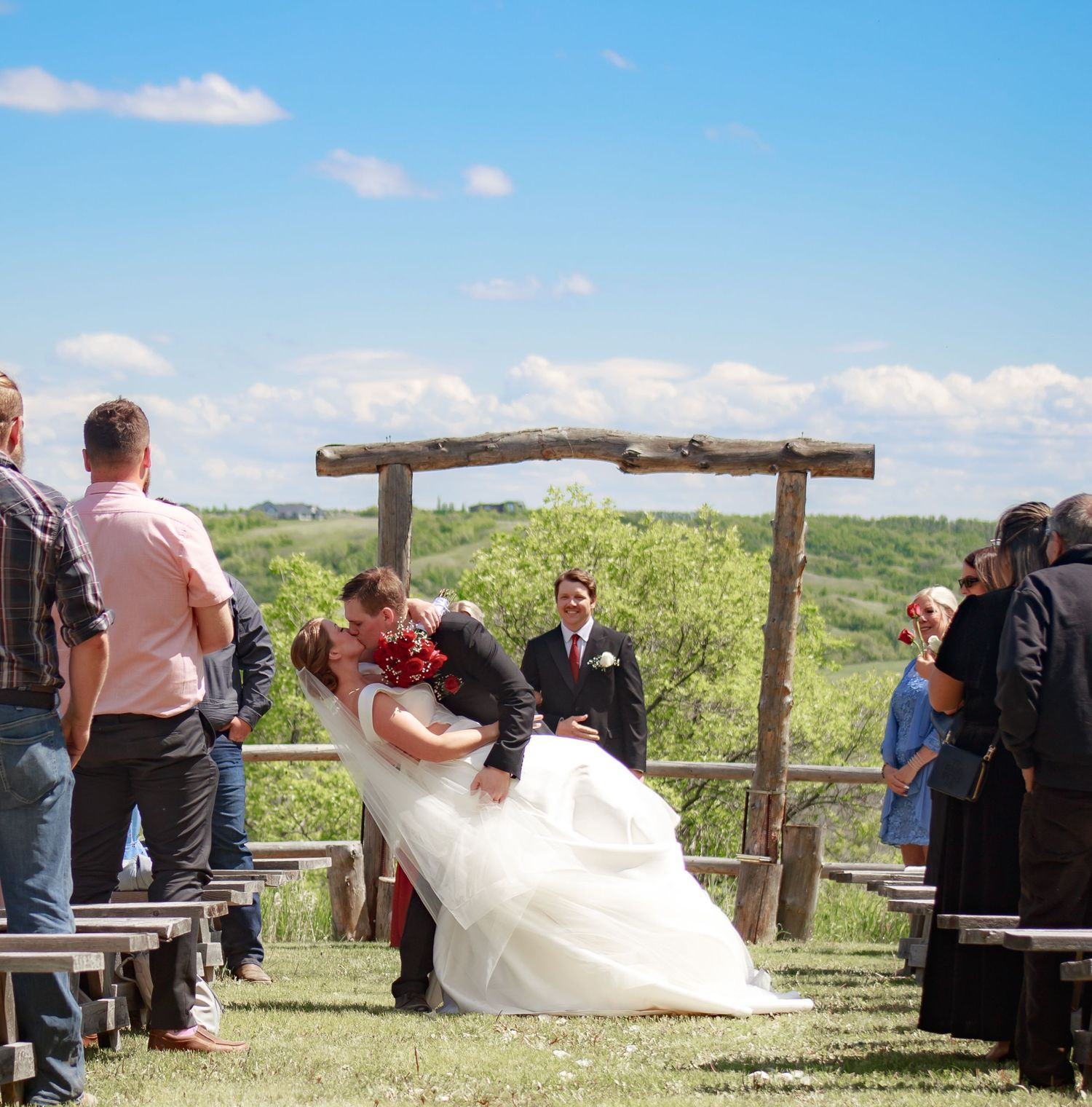 Rustic outdoor wedding ceremony setup with wooden arch against blue sky and rolling hills.